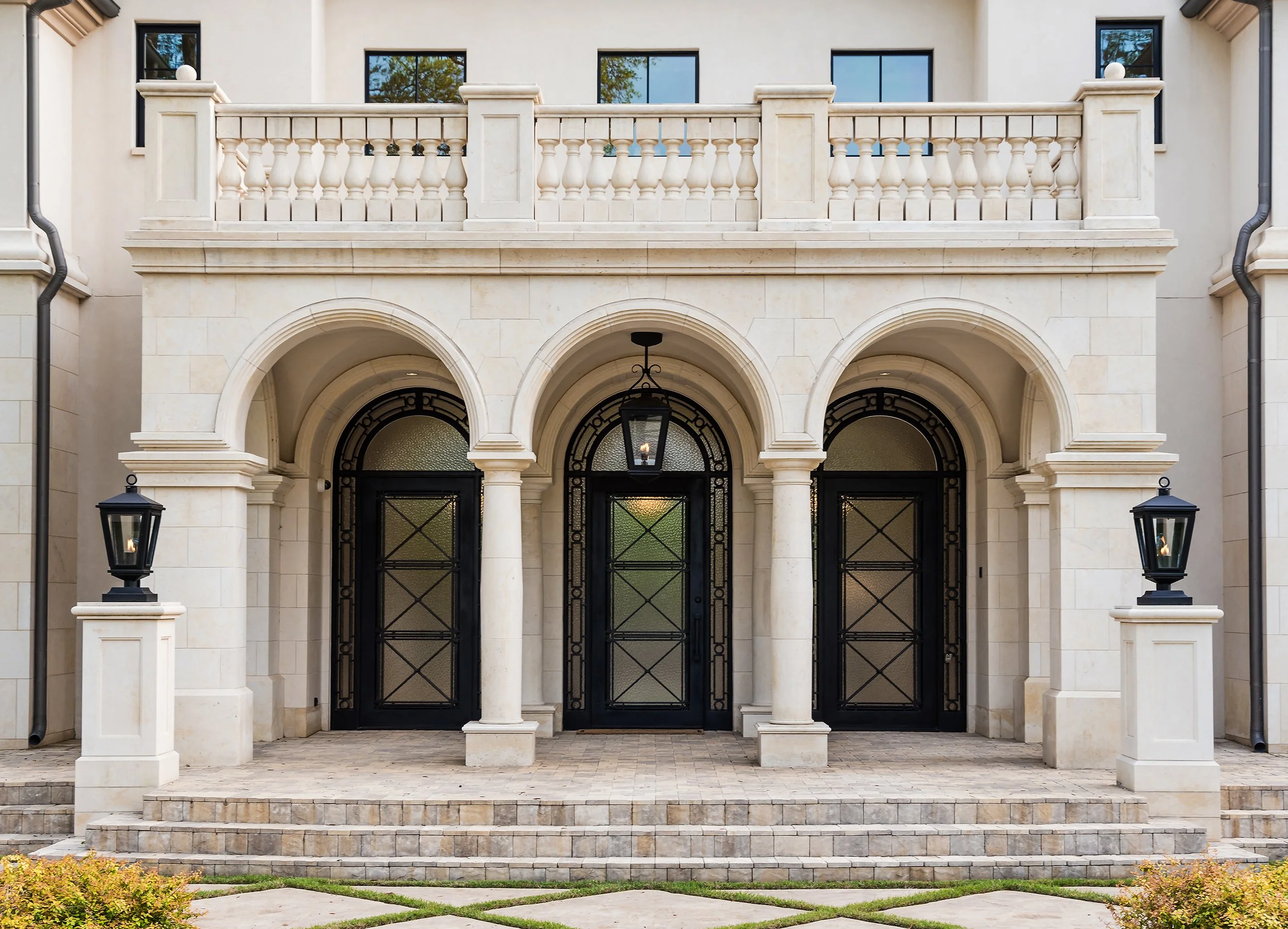 Front view of a house entrance featuring arched doorway, black double doors with decorative metalwork, a lantern hanging from the ceiling, and two lantern-style light fixtures on stone pillars flanking the steps. There is a balcony above the entrance with stone balustrades and three windows behind it. The construction style is elegant and classical with light-colored stone and stucco exterior.
