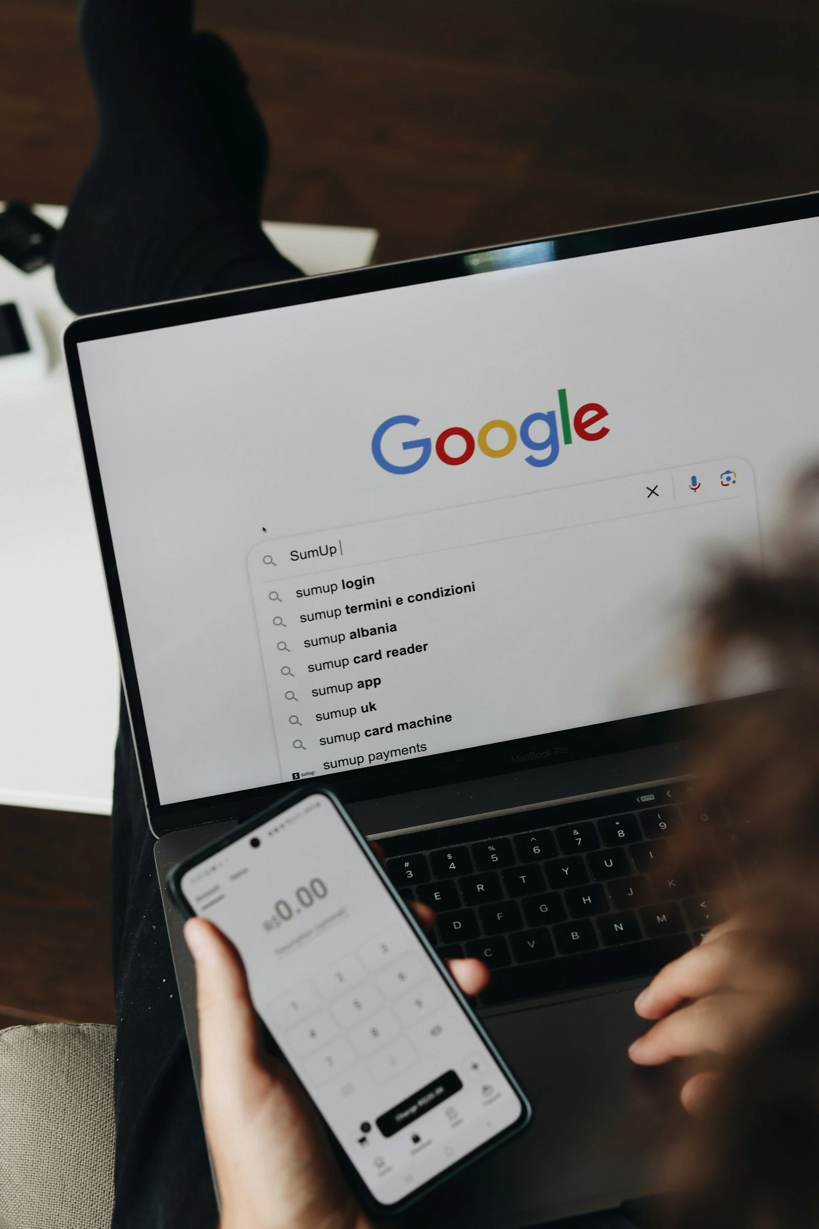 Person using a laptop displaying Google search results for "SumUp" alongside a smartphone showing a payment interface, illustrating digital marketing and online payment solutions.