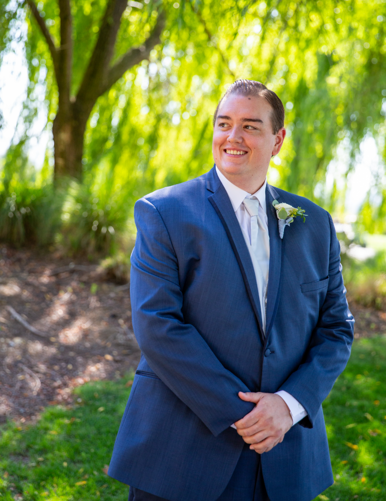 Man in a blue suit with a boutonniere, smiling outdoors in a lush green setting, representing a professional image relevant to digital marketing consultancy.