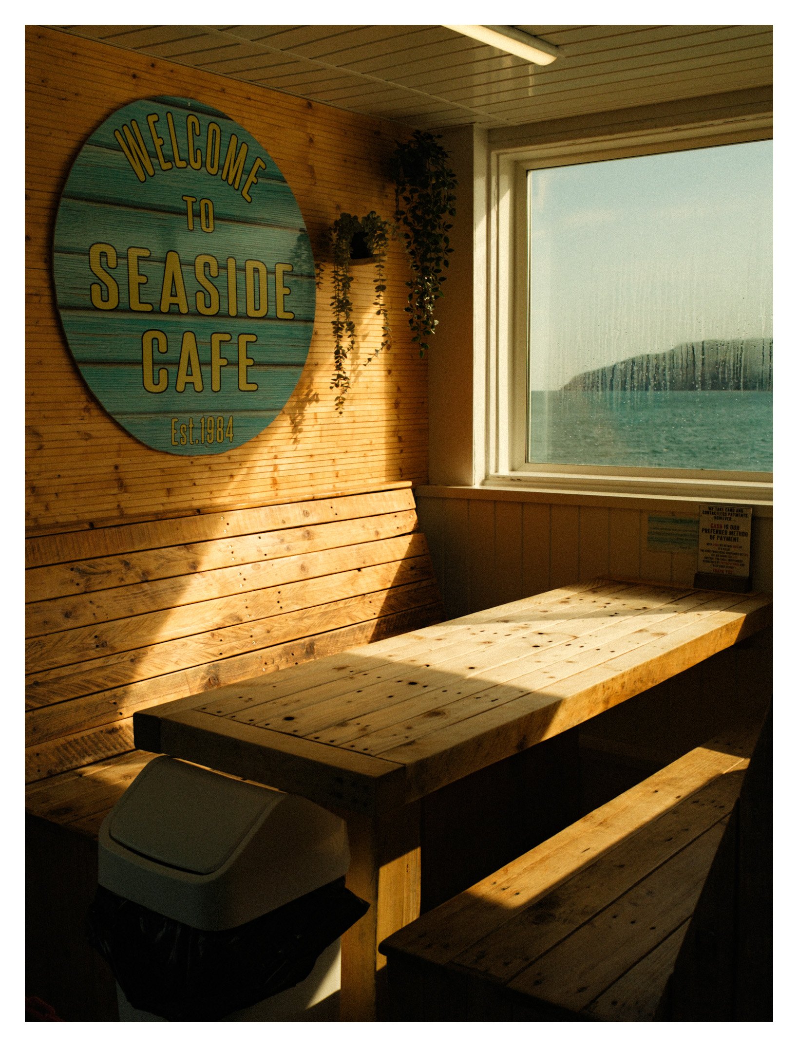 Indoor seating area with wooden bench and tables, a large window with ocean view, and a 'Welcome to Seaside Cafe' sign on the wall.