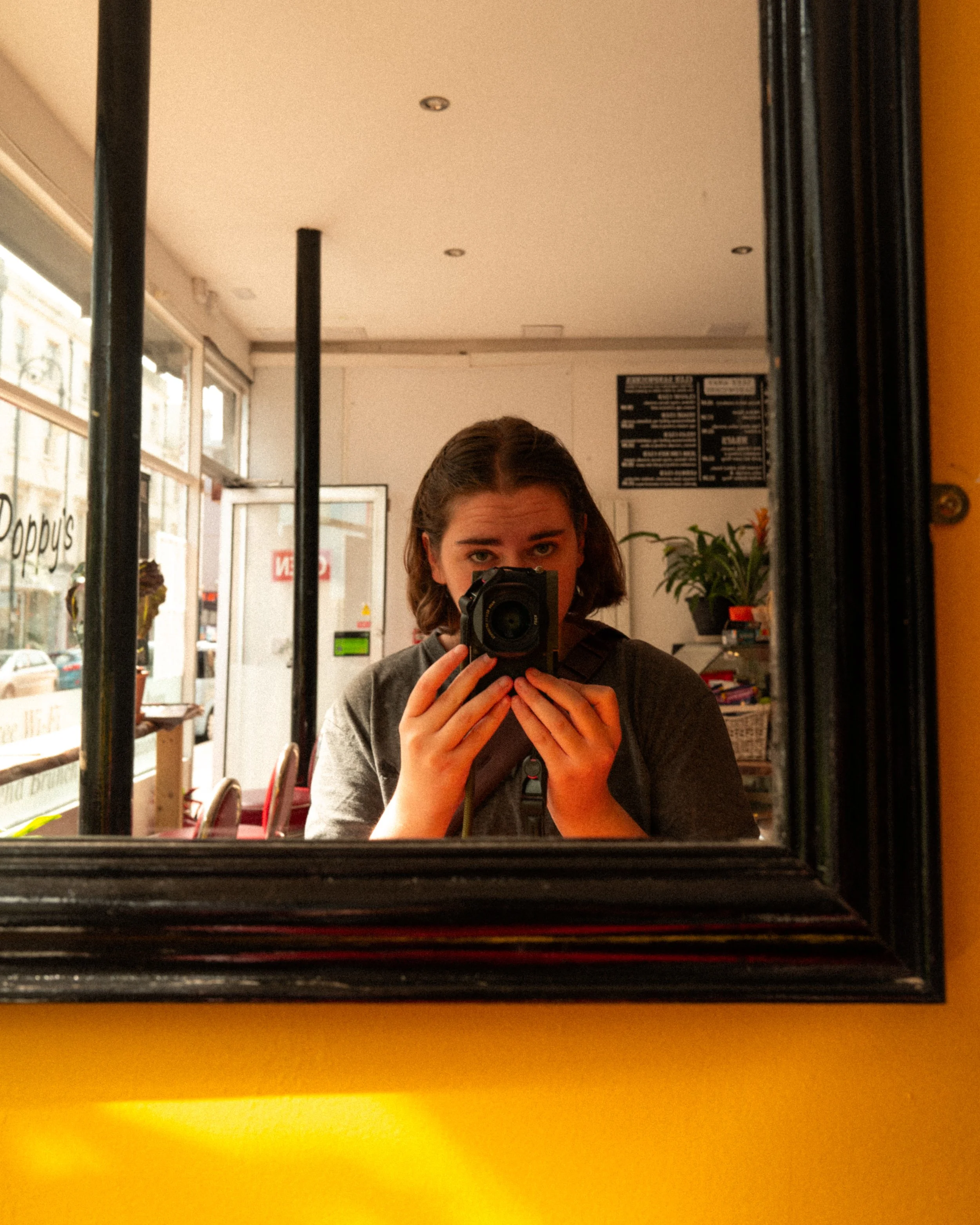 A woman taking a mirror selfie inside a restaurant or cafe, with a camera covering her face, in front of a black-framed mirror. The background shows a menu on the wall, some plants, and the entrance door.