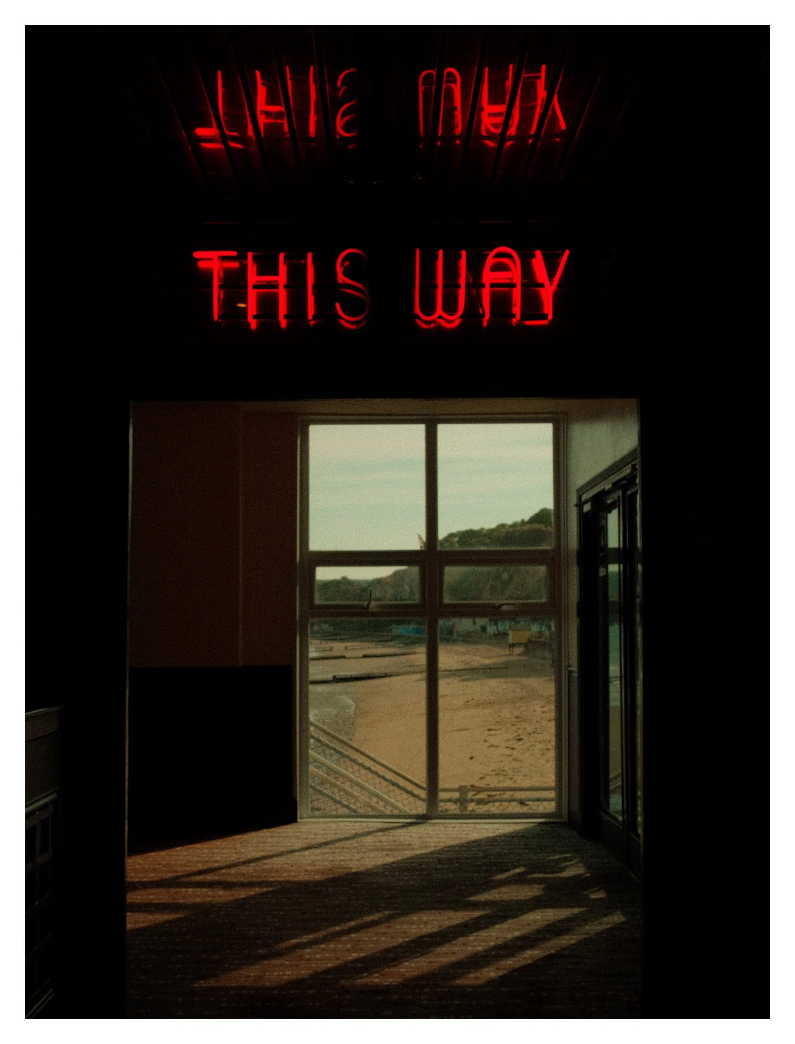 A red neon sign reads 'THIS WAY' above a doorway, with an outside view of a road, landscape, and cloudy sky visible through a glass door and window.