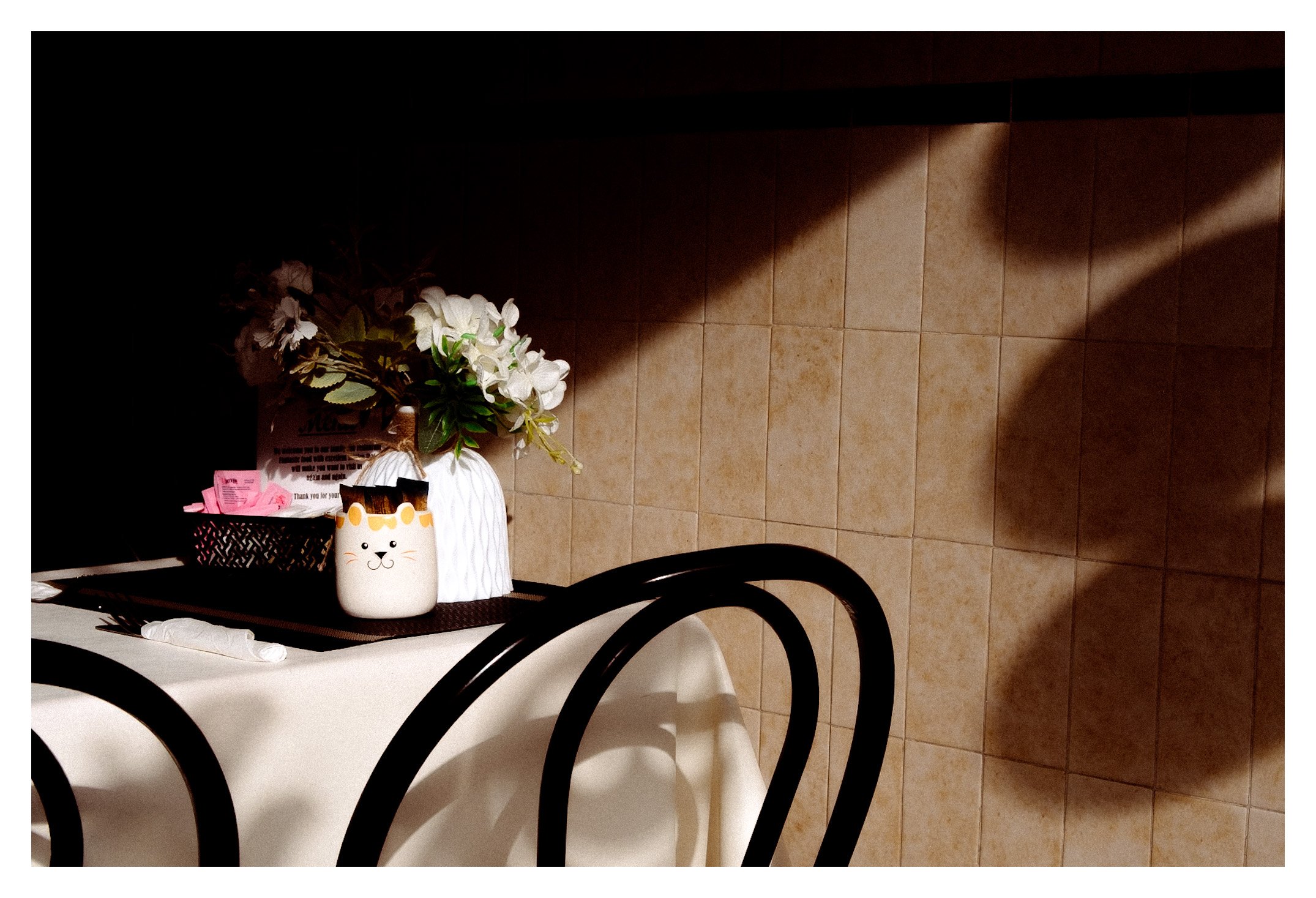 A table in a restaurant or cafe with a black metal-framed chair in the foreground, a potted white flower, a basket with pink papers, and a white paper bag with a cat face illustration. The background includes beige tiled walls with shadows cast acros