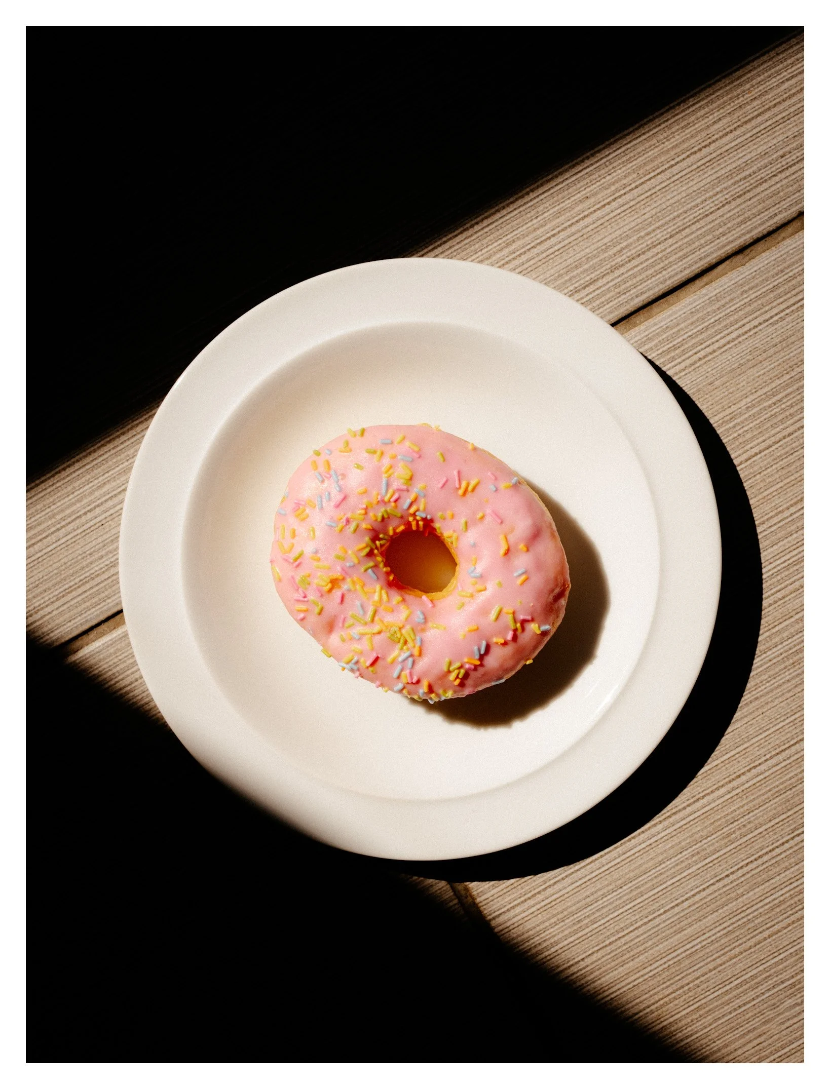 A pink frosted donut with colorful sprinkles on a white plate, placed on a wooden table with a dark background.