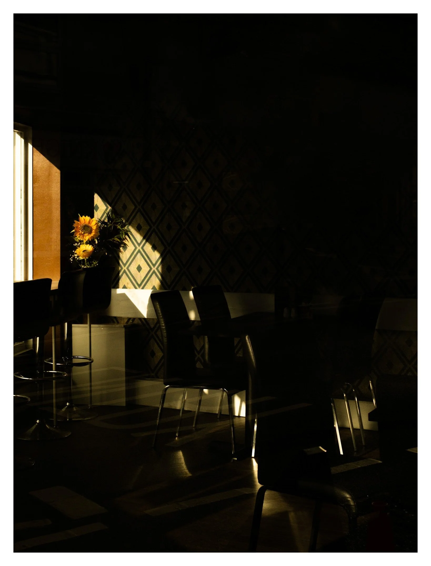 Sunlight streaming into a dark room with a window, illuminating a vase of sunflowers on a table and a patterned wall in the background.