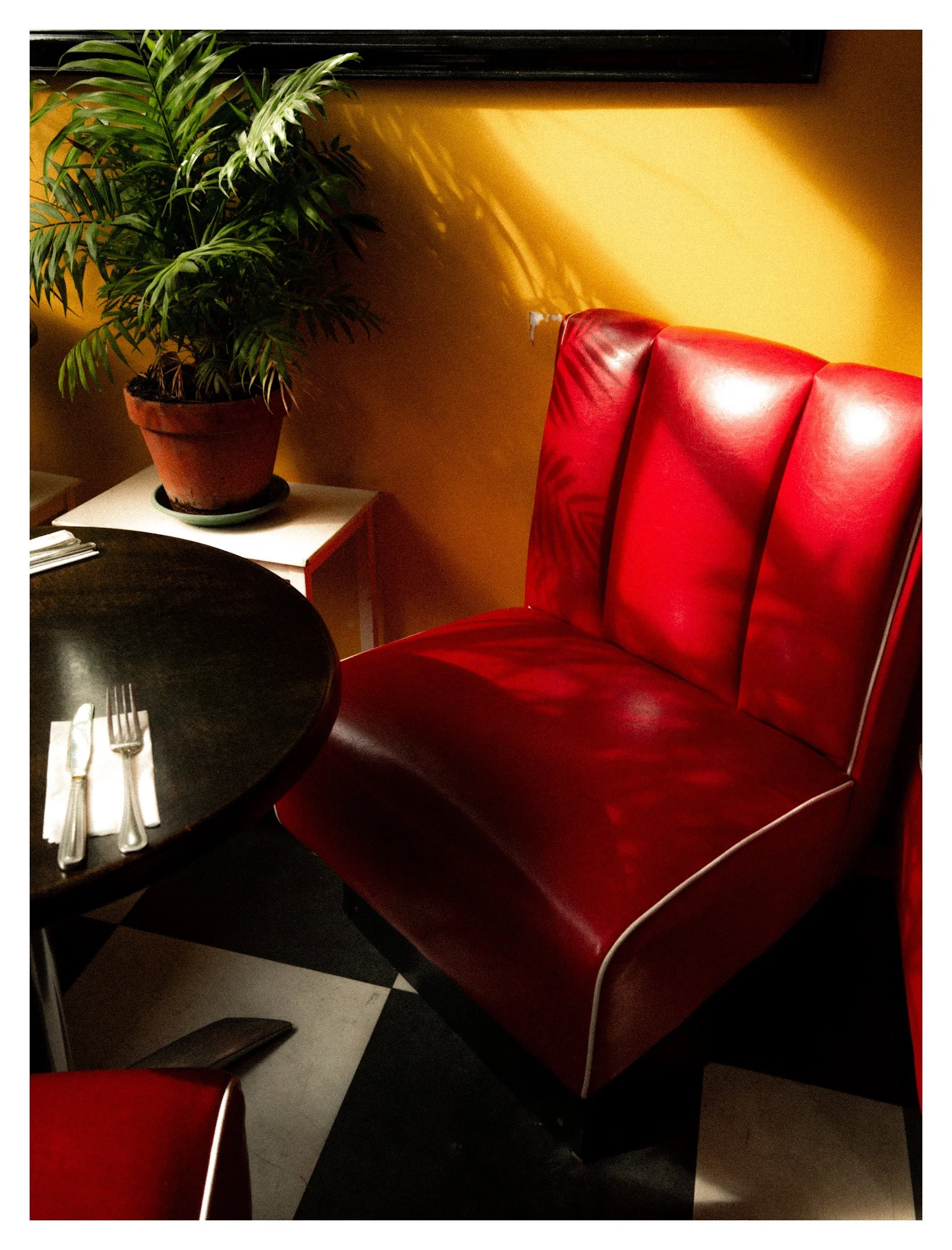 A red leather booth seat in a restaurant, a potted plant on a nearby white table, and a partially visible black table with silverware in the foreground. A black frame is mounted on a yellow wall behind the plant.