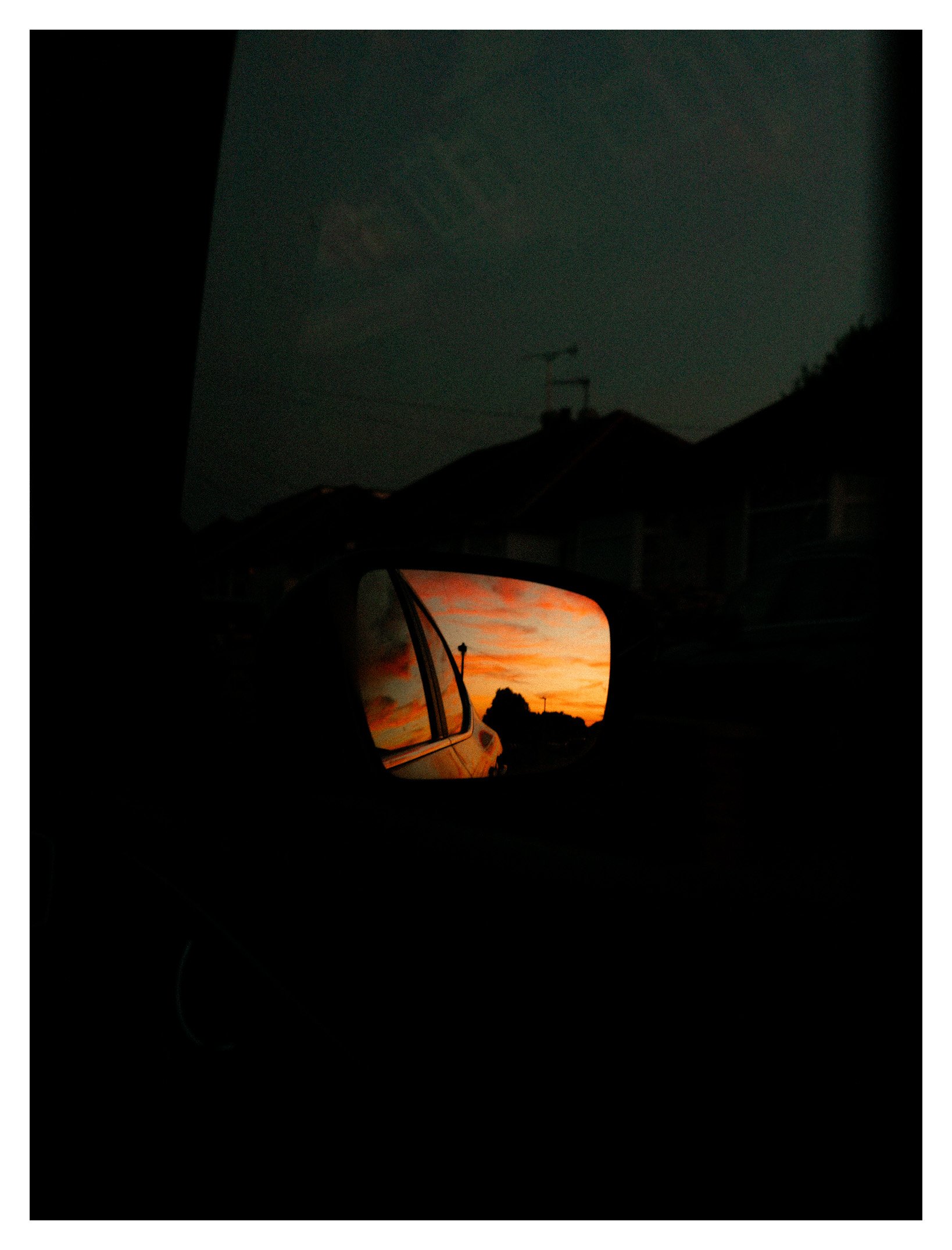 A sunset view seen through a car's side mirror with an orange and pink sky and silhouettes of trees in the distance.