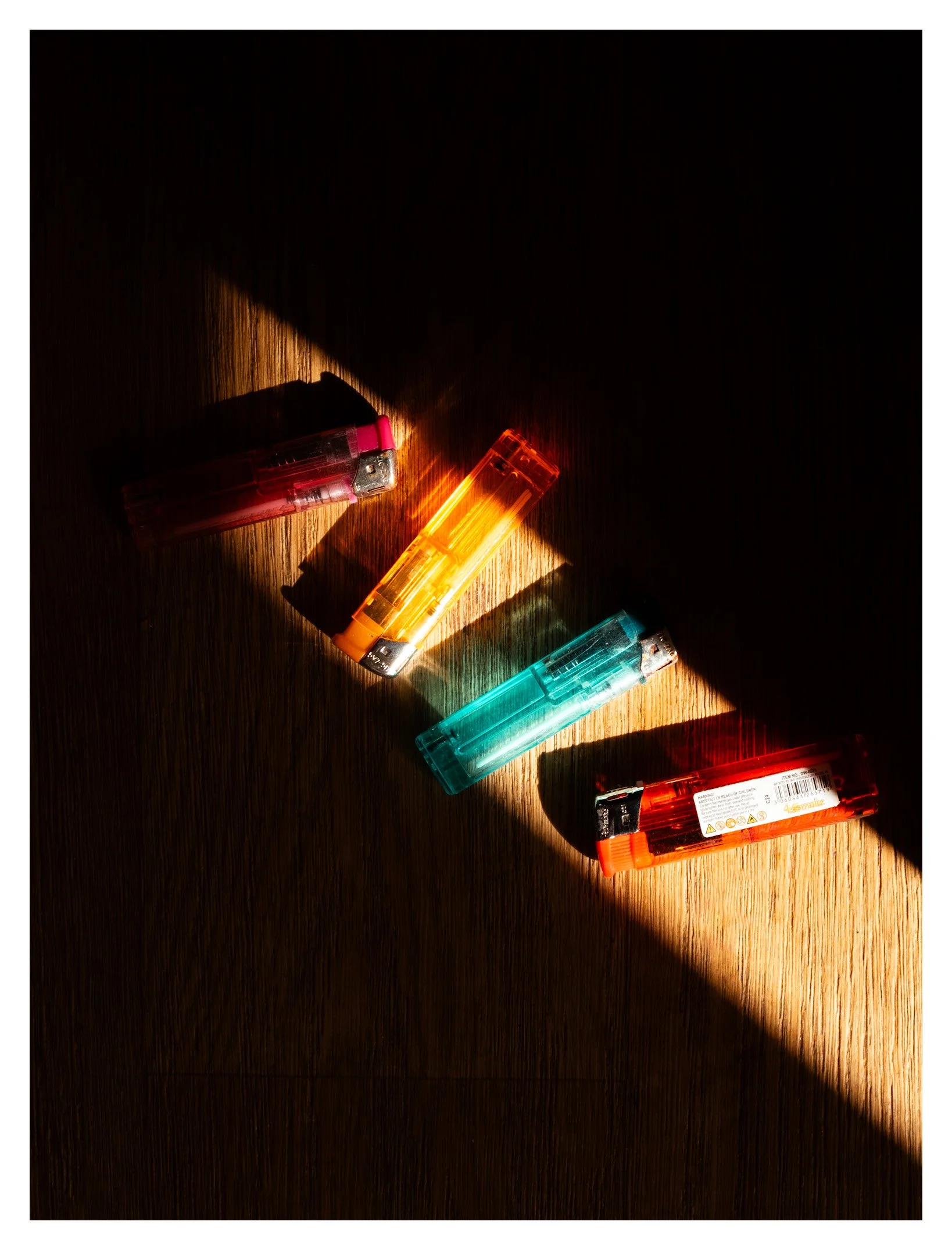 Colorful disposable lighters lying on a wooden surface with sunlight casting shadows.