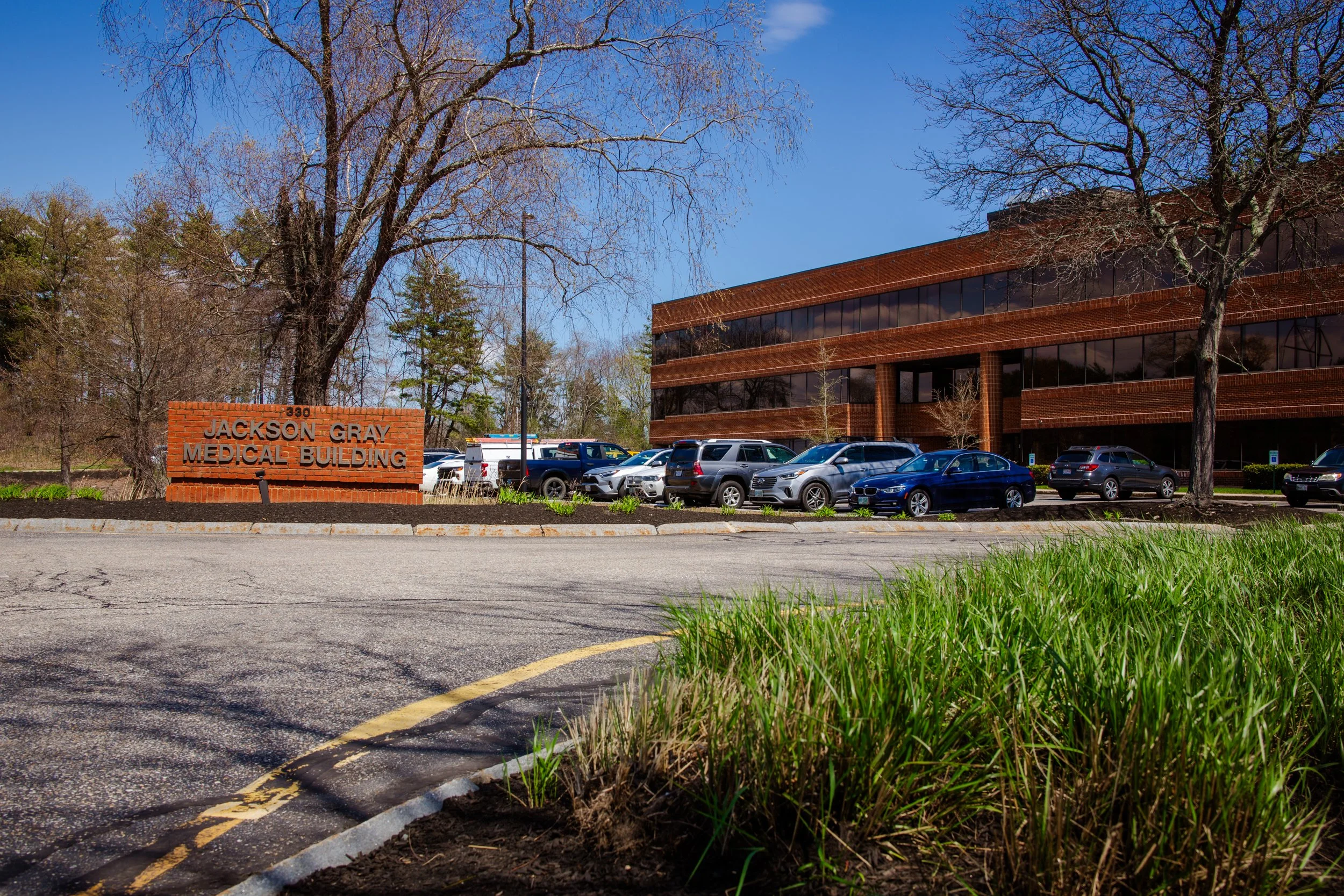Photo of the Jackson Gray Medical Building, a large red brick office building with reflective windows, parked cars in front, trees on the property, and a clear blue sky in the background.