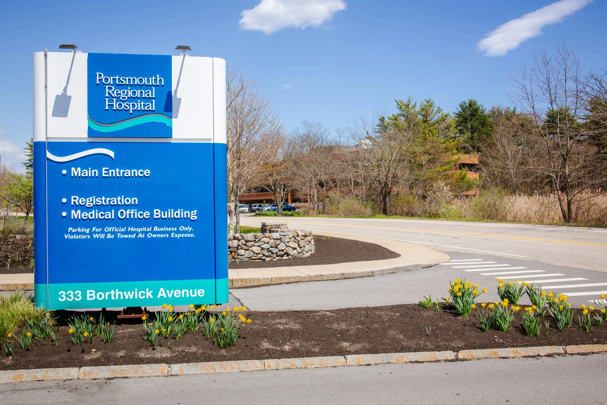 Blue sign for Portsmouth Regional Hospital at 333 Borthwick Avenue, with parking area, trees, and a building in the background on a clear day.