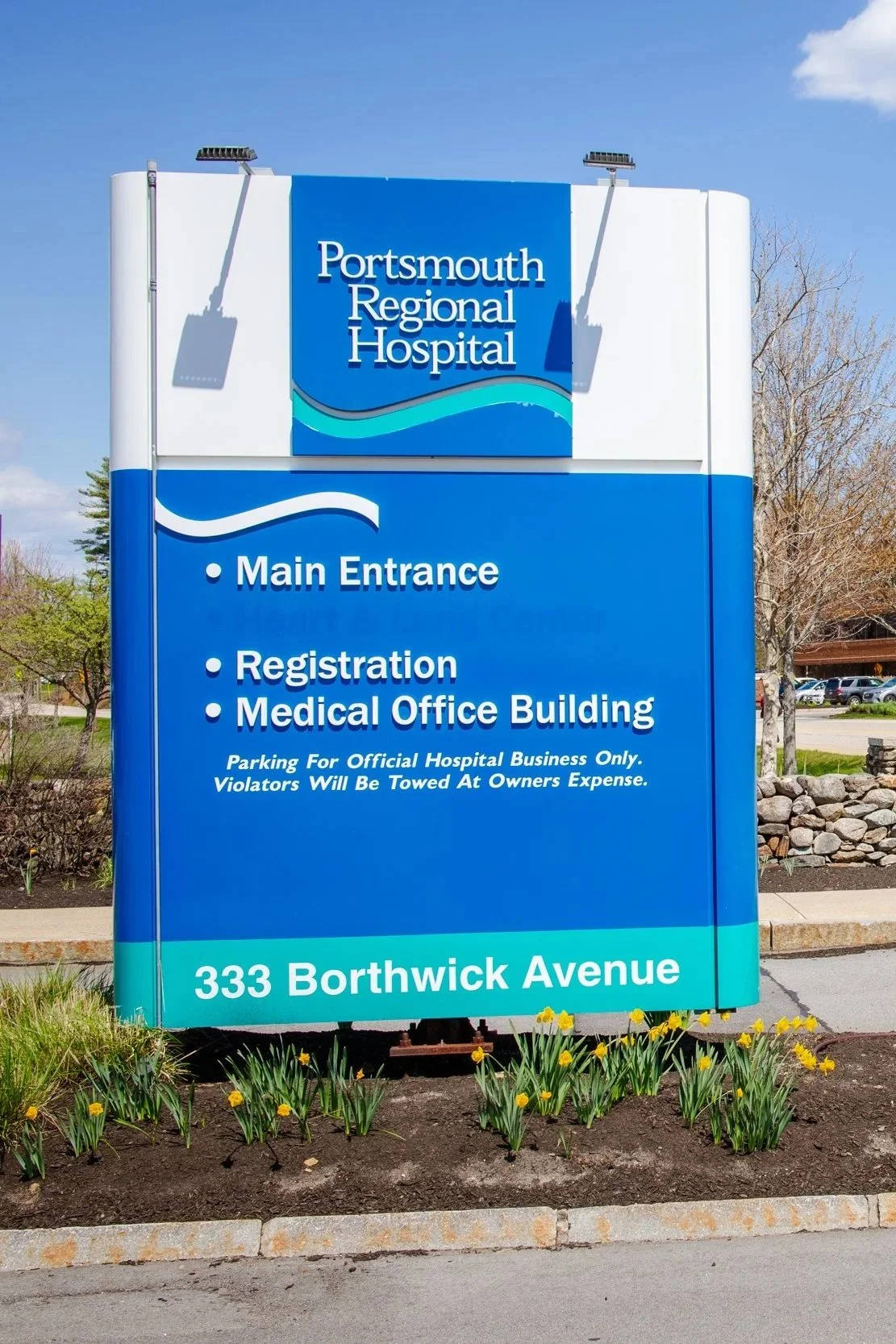 Blue sign for Portsmouth Regional Hospital with directions to main entrance, registration, and medical office building, located at 333 Borthwick Avenue, with spring flowers in the foreground.