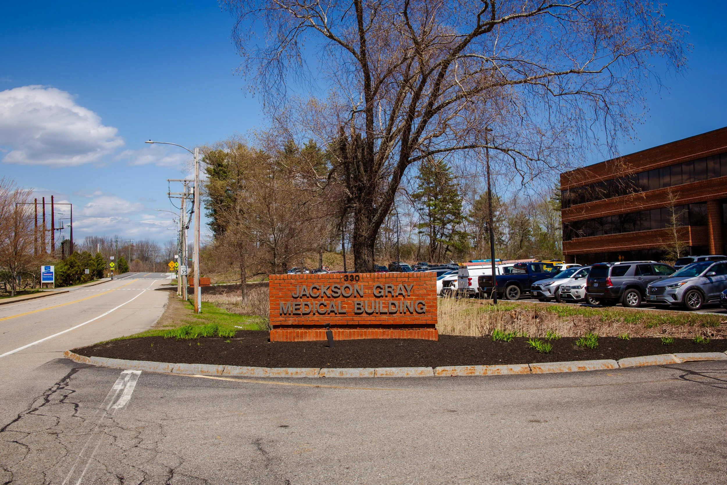 A parking lot in front of Jackson Gray Medical Building with a brick sign, trees, parked cars, and a building with reflective windows under a blue sky with clouds.