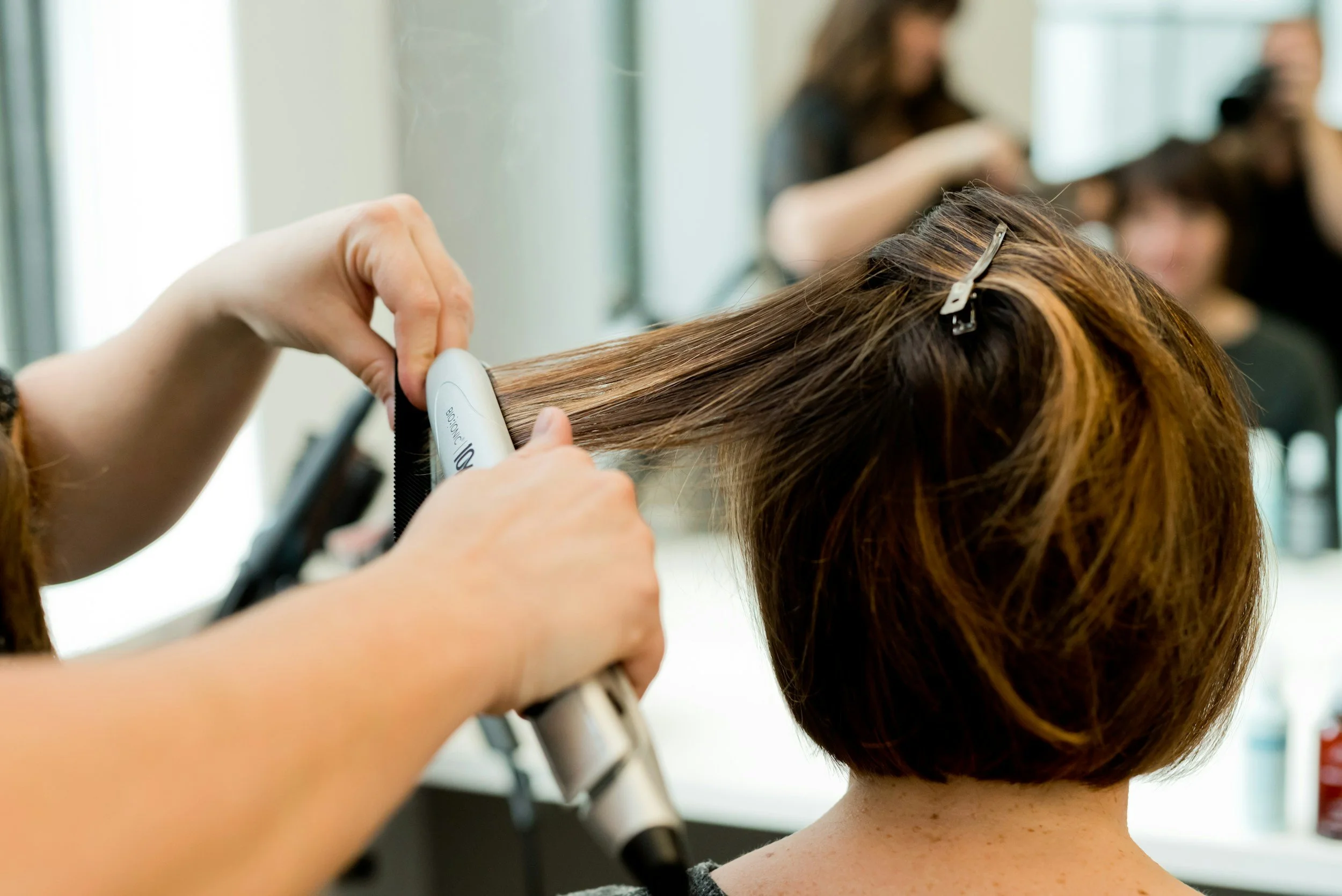 A hairstylist using a flat iron to straighten a woman's short, reddish-brown hair in a hair salon.