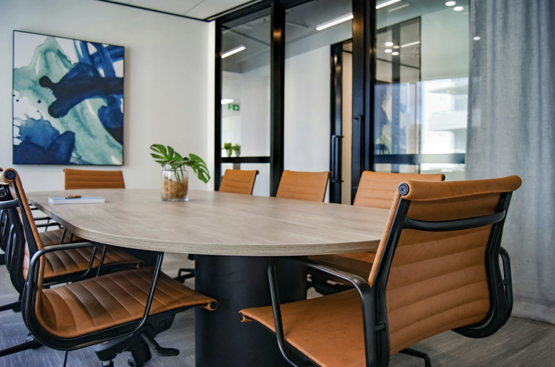 A conference room with a wooden table surrounded by brown leather chairs, a potted plant in the center, a large abstract painting on the white wall, and glass walls with a door leading to another room.