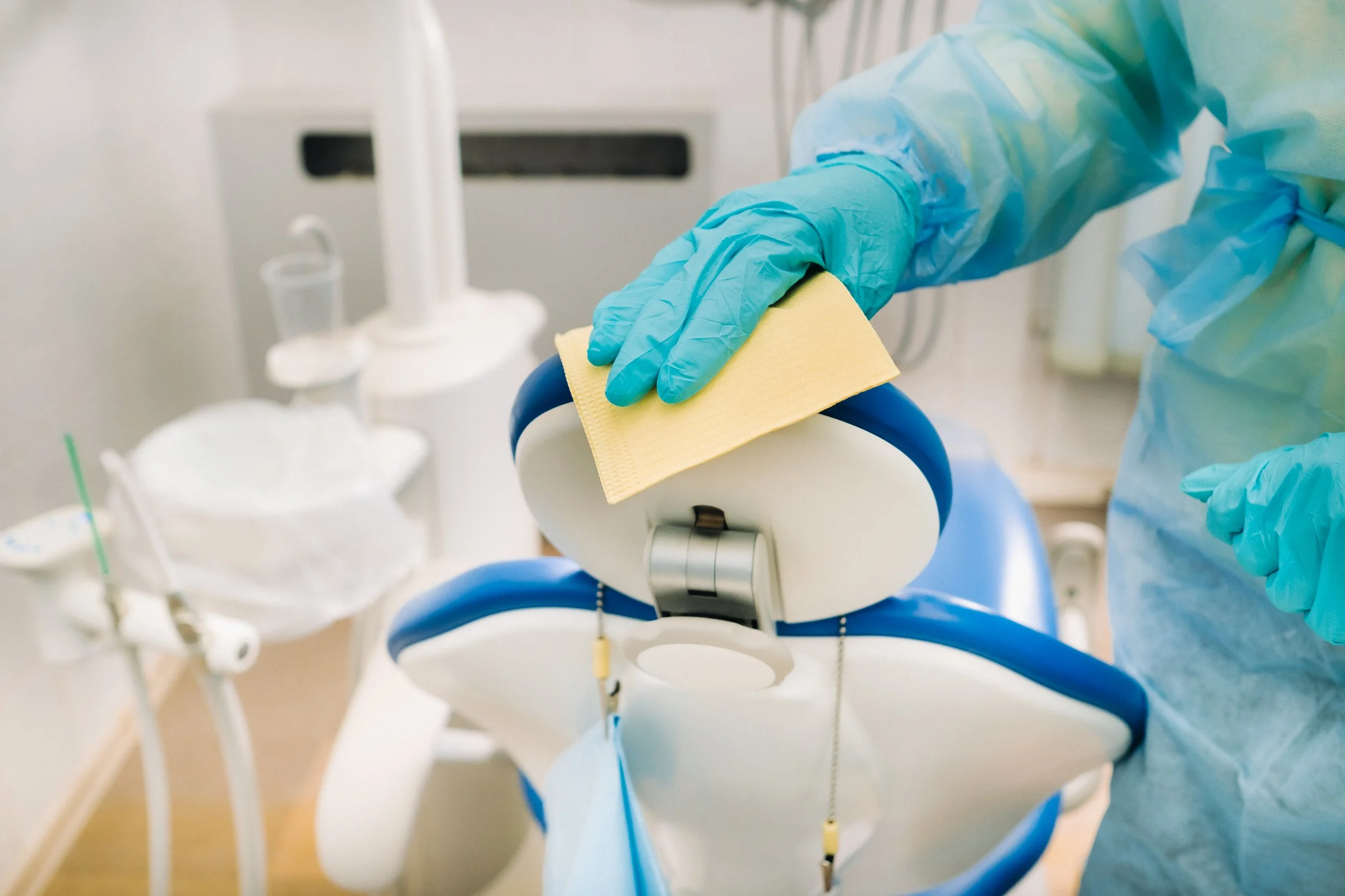Healthcare worker disinfecting a dental chair with a yellow cloth in medical attire and blue gloves.