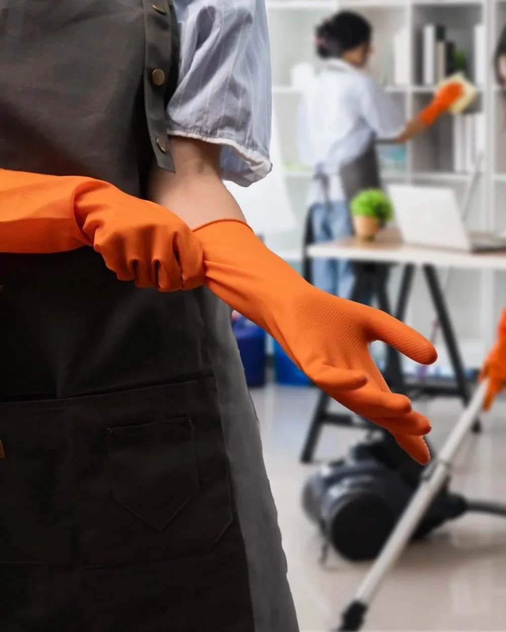 Person wearing orange cleaning gloves, in a cleaning or maintenance setting, with a person in the background cleaning a white shelf.