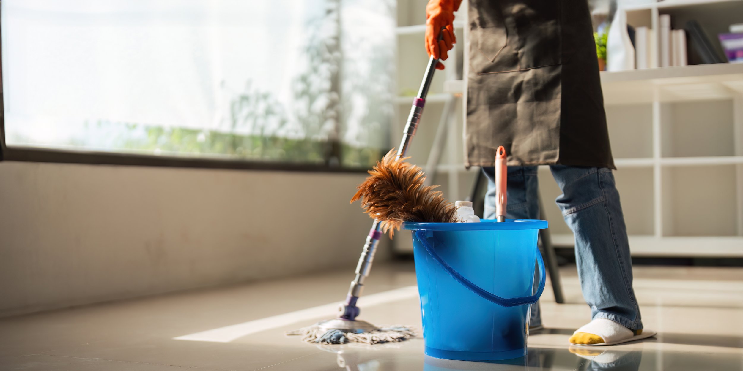 Person mopping floor with bucket and cleaning supplies in a bright room.