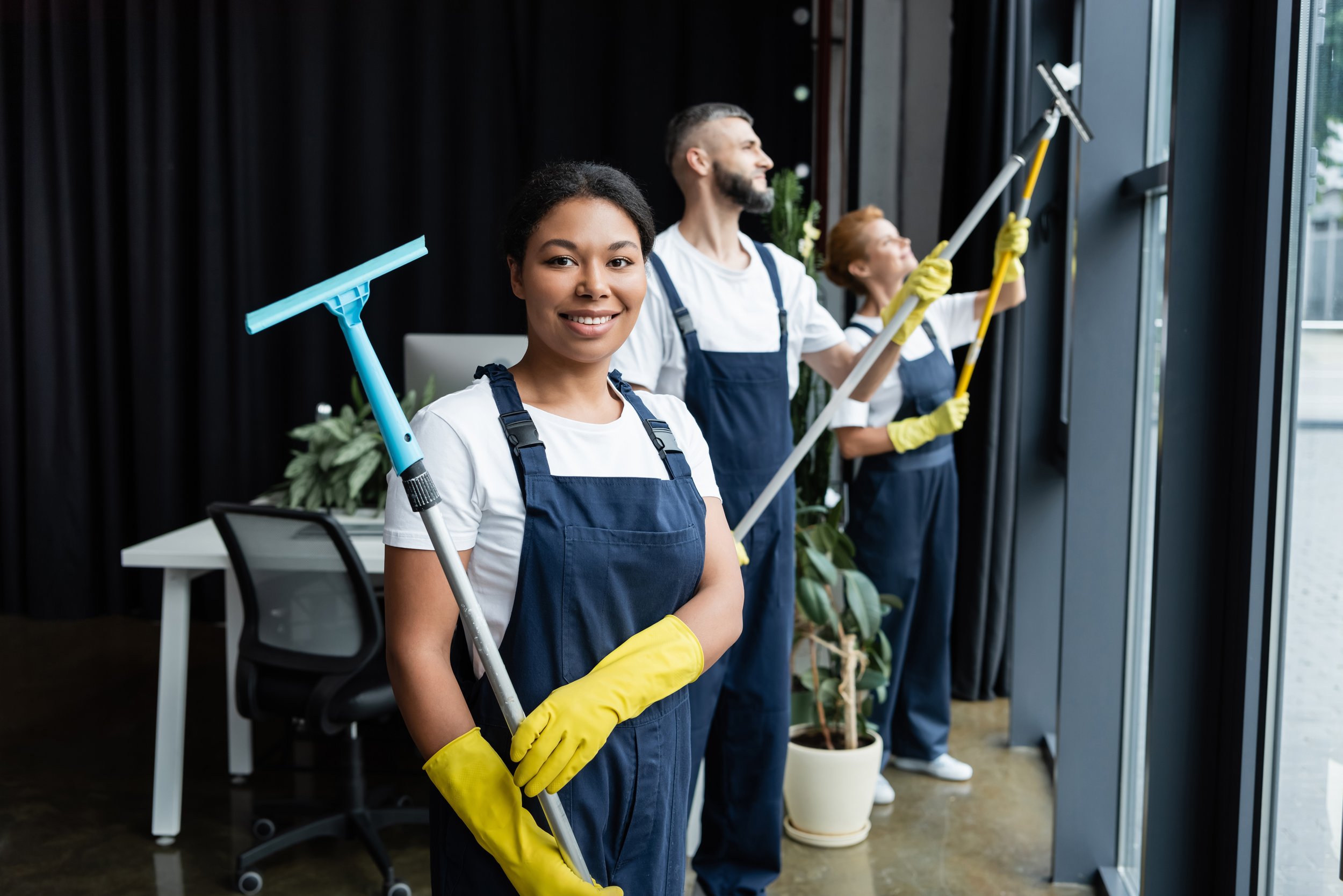 Three diverse people cleaning large windows with squeegees and scrub brushes wearing yellow rubber gloves and aprons.