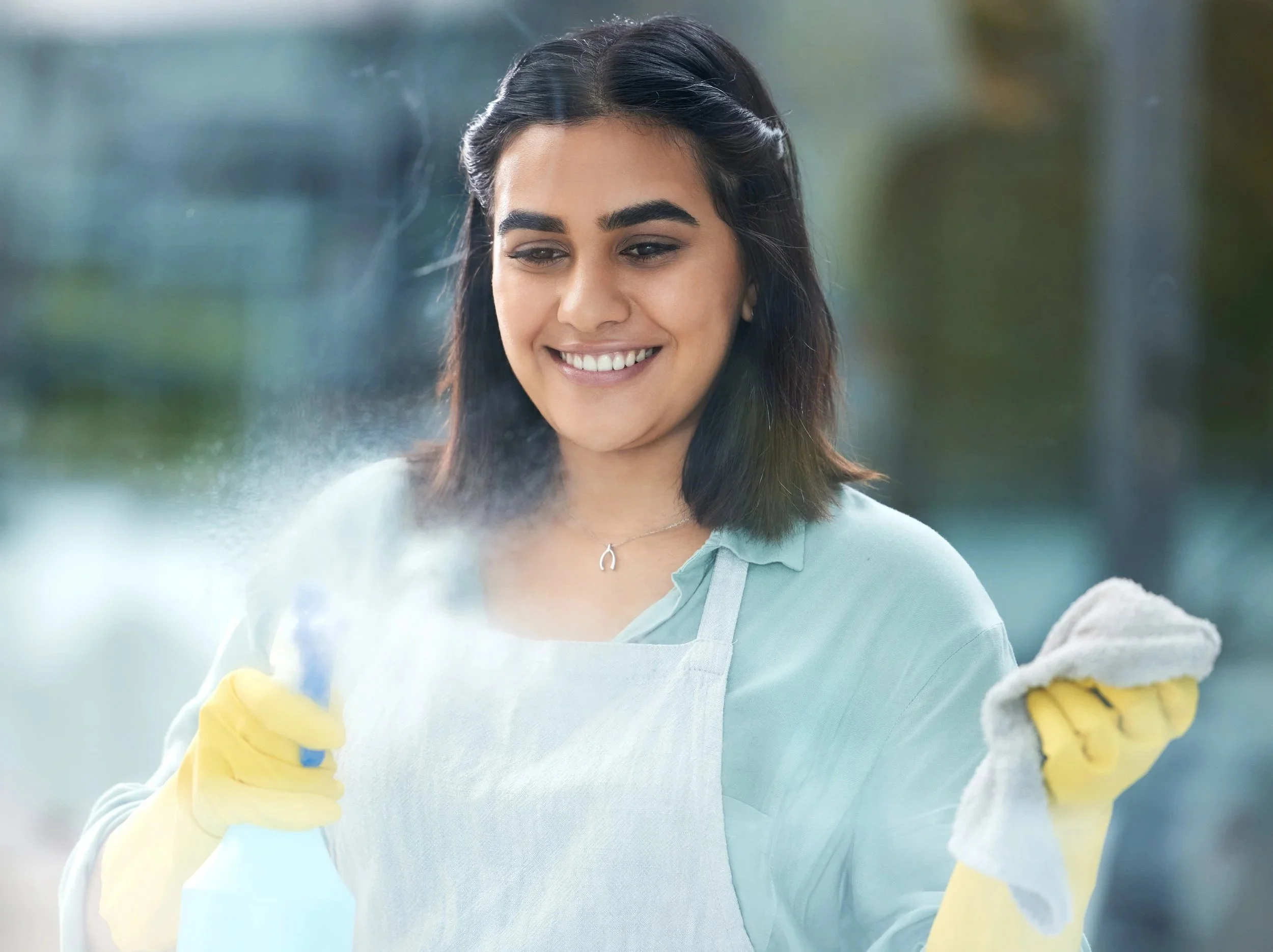 A woman wearing yellow gloves and a light blue shirt with a collar, smiling while cleaning a window with a cloth.