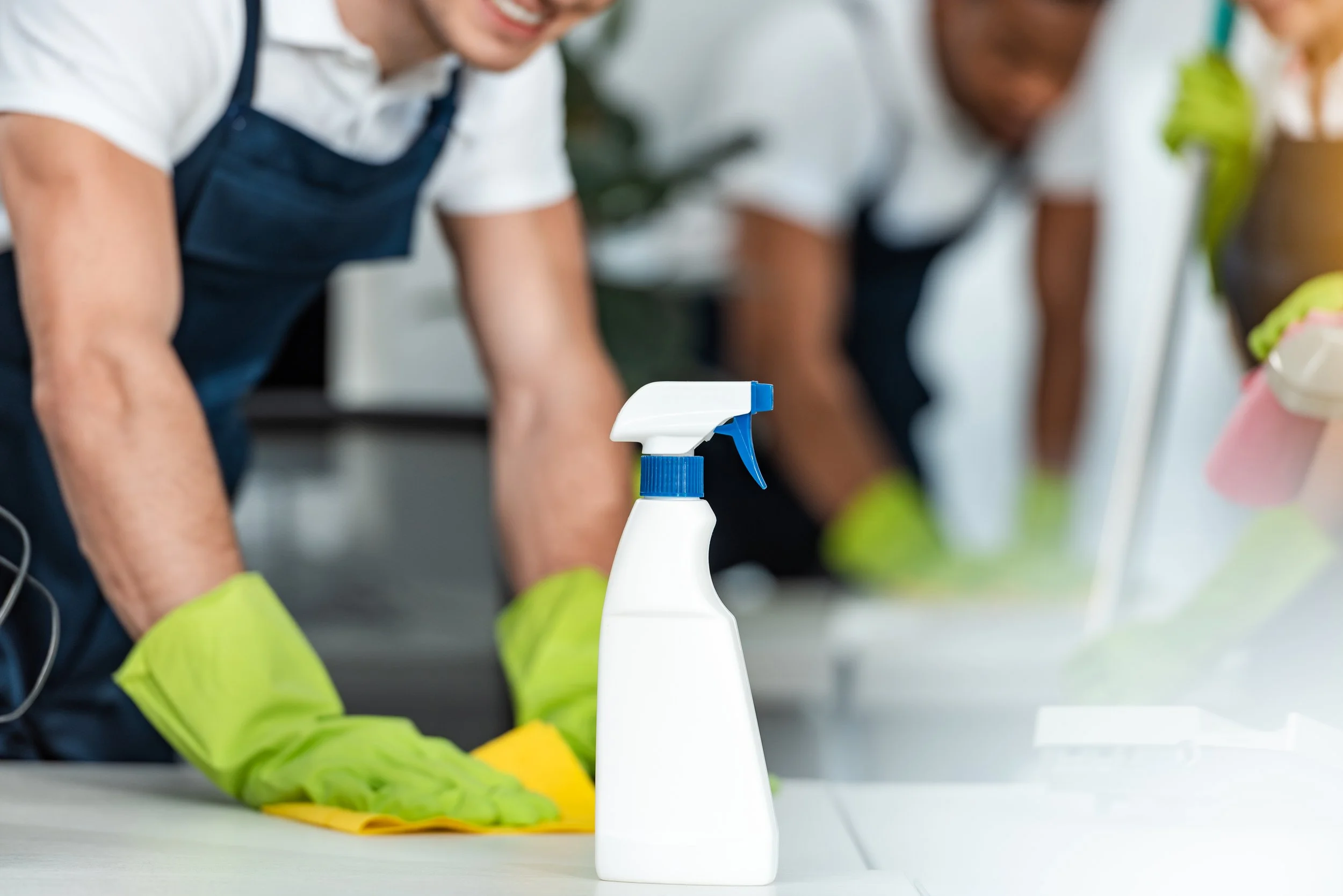 Cleaning spray bottle on a table with people wearing gloves and aprons cleaning in the background
