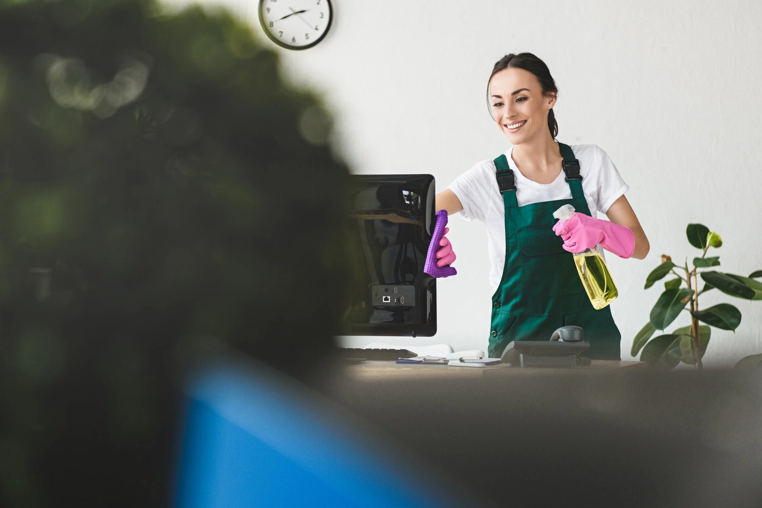 A woman wearing pink gloves and a green apron cleaning a computer monitor with a spray bottle and cloth in an office setting.