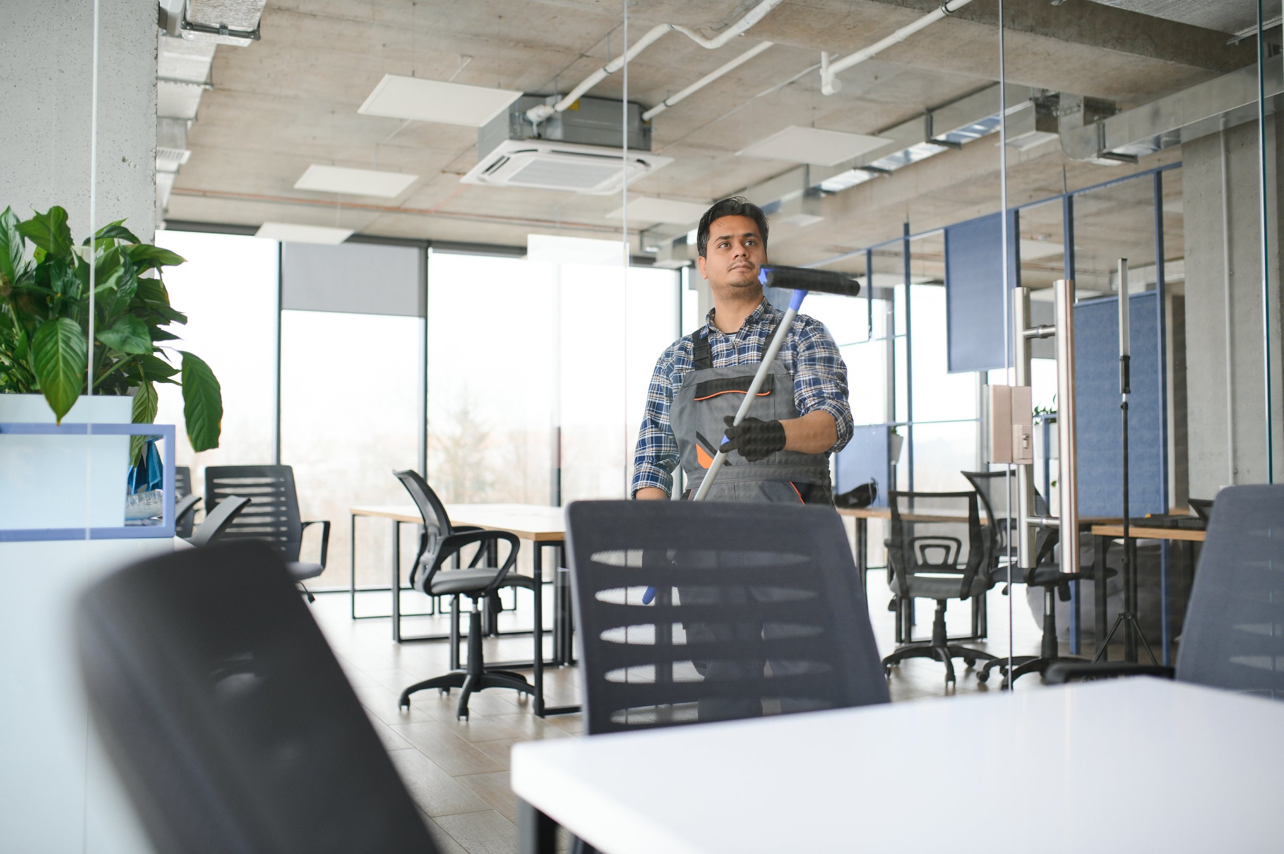 A man in a plaid shirt and work apron, wearing gloves, is using a squeegee to clean a window in an office space.