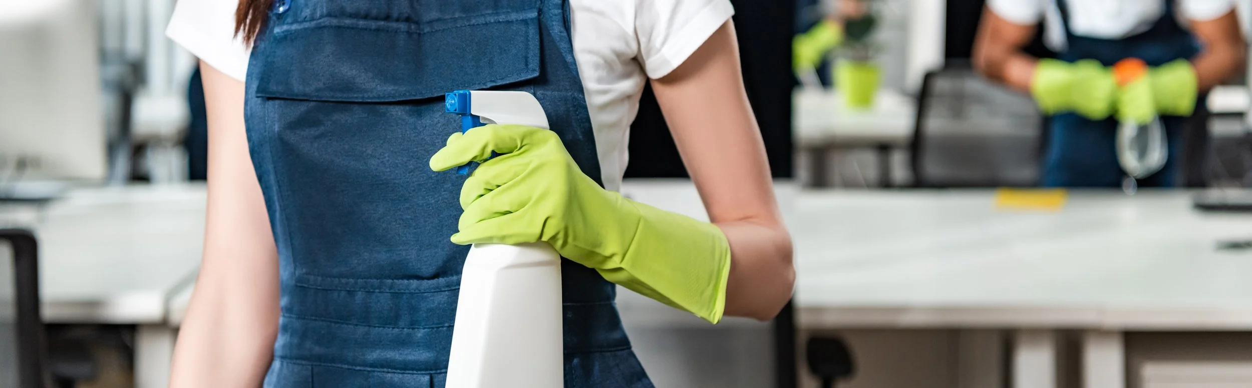 Person wearing a blue apron and bright green gloves holding a spray bottle in a cleaning or disinfecting setting with other people in the background.