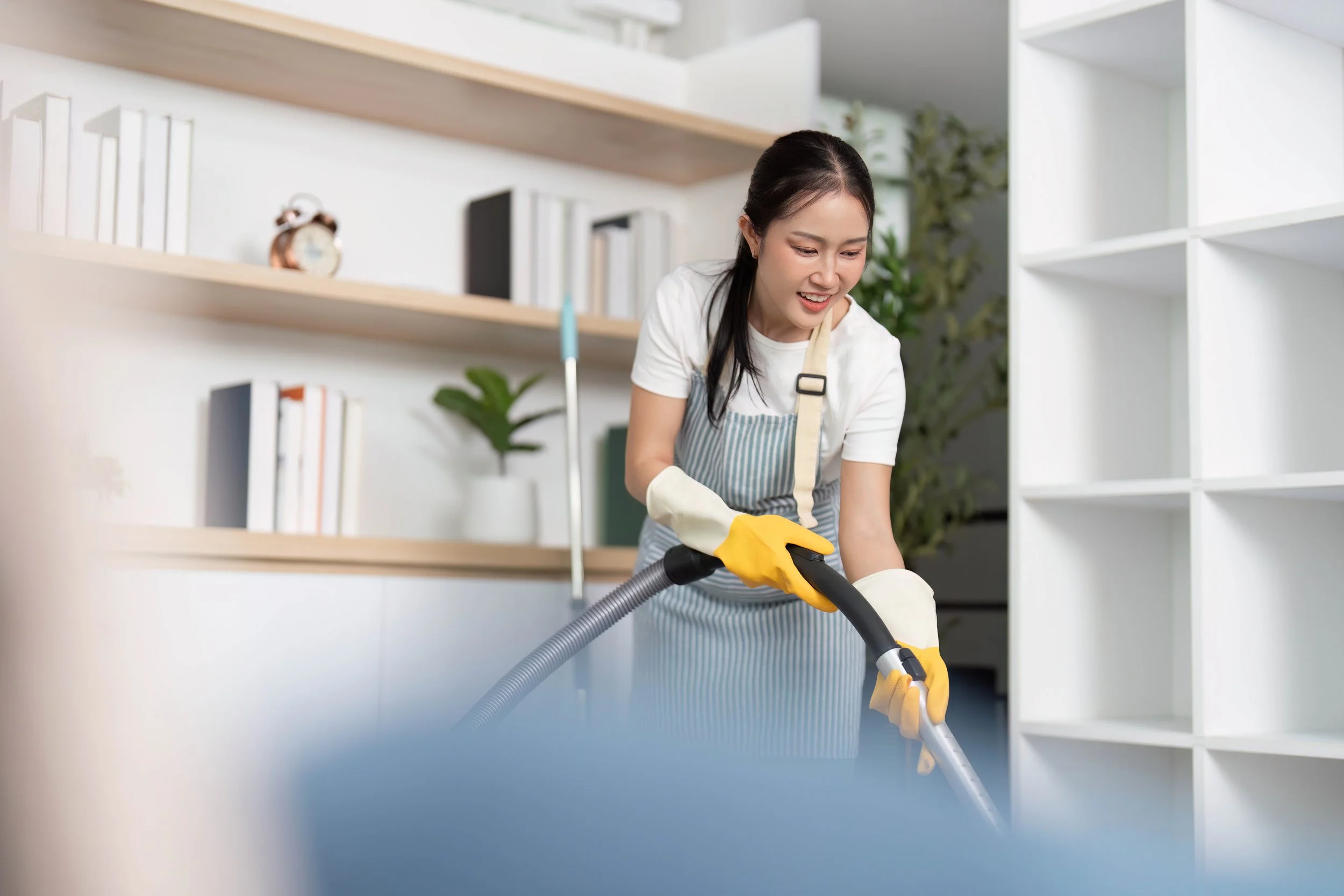 A woman wearing yellow cleaning gloves and an apron is vacuuming a room with a modern vacuum cleaner, smiling as she works.