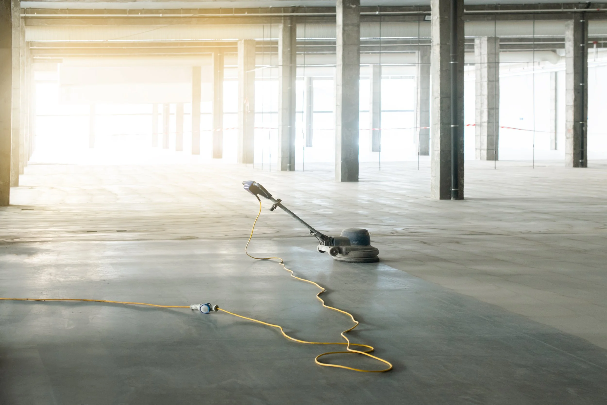An indoor construction site with concrete columns and large windows, featuring a floor polishing machine with a yellow cord.