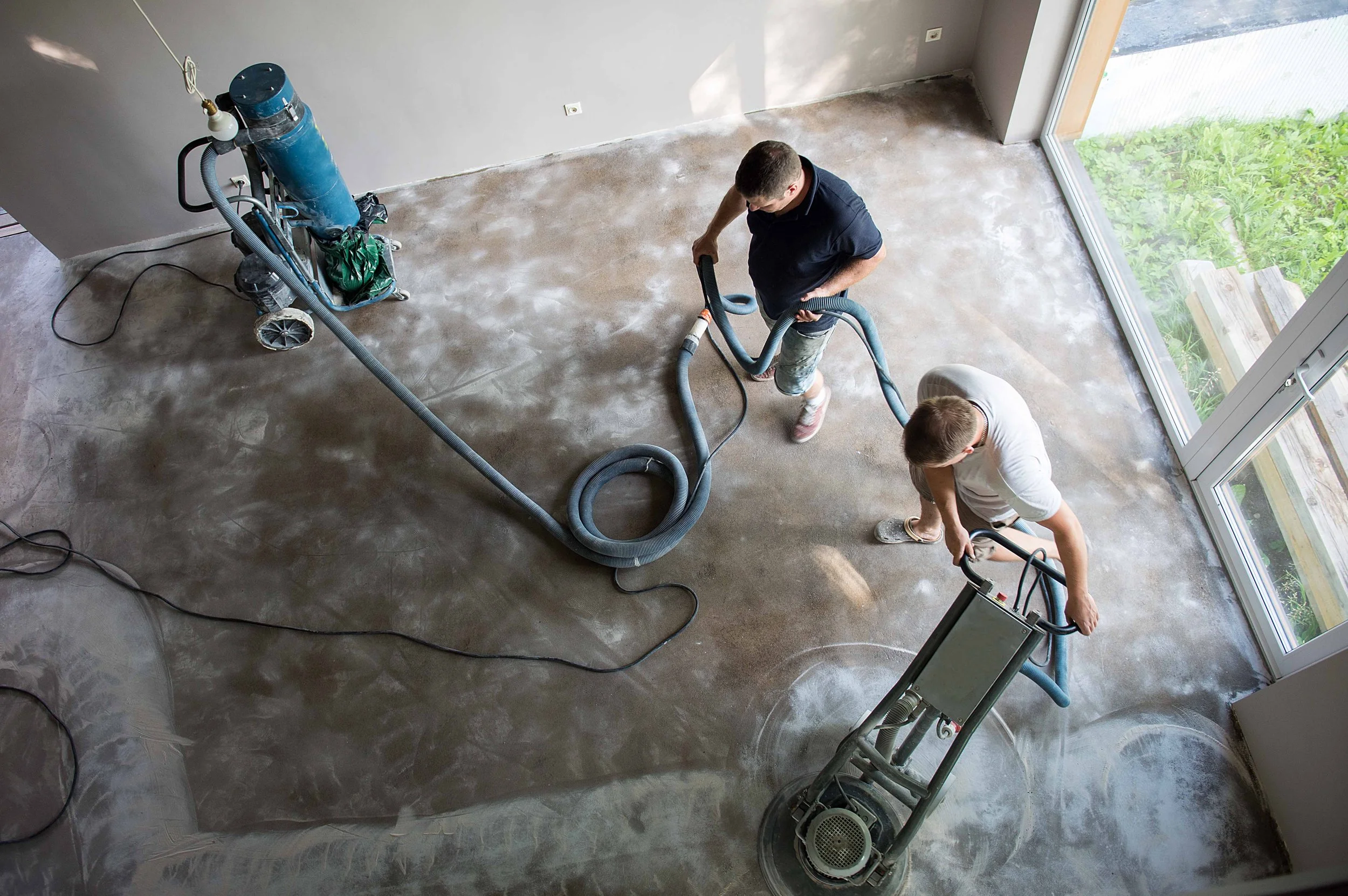 Two workers are vacuuming a concrete floor in a room with large windows, with construction equipment nearby.