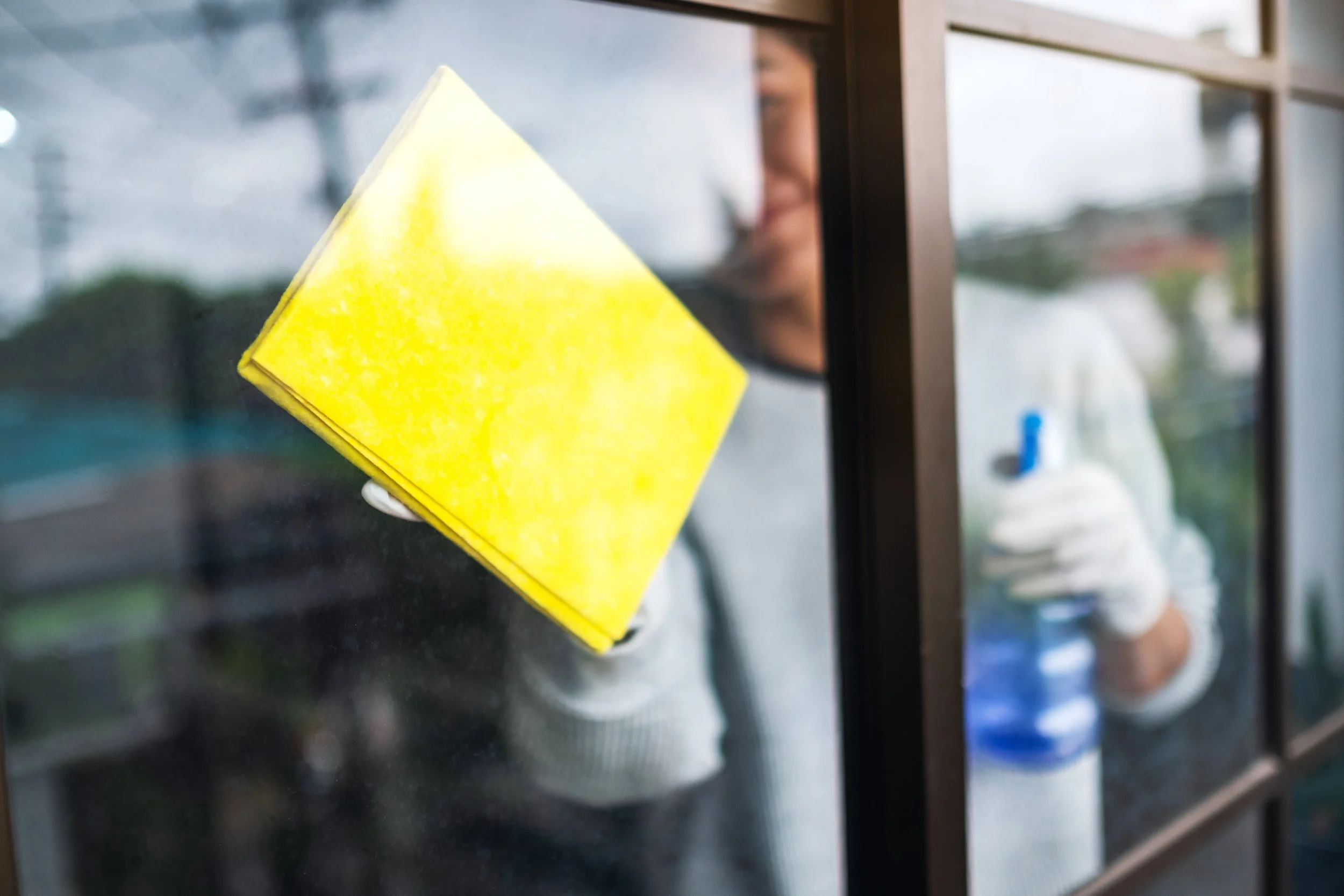 Person cleaning glass door with yellow cloth while wearing gloves, holding spray bottle.