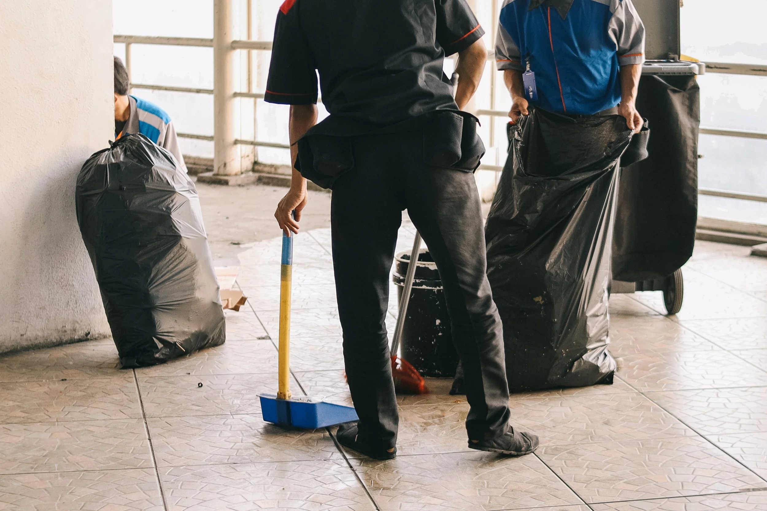 Two sanitation workers, one in black and one in blue uniforms, working on a tiled sidewalk with garbage bags and a trash bin during daytime.