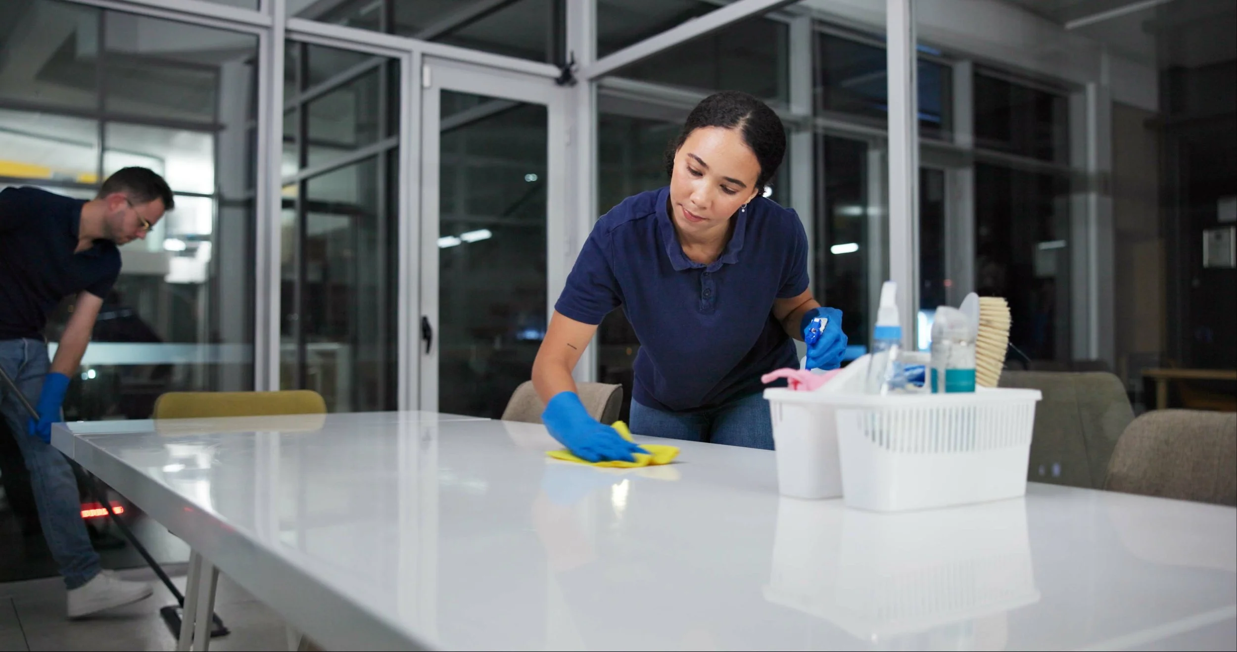 Two people cleaning a conference room table at night, wearing blue gloves and using cleaning supplies.