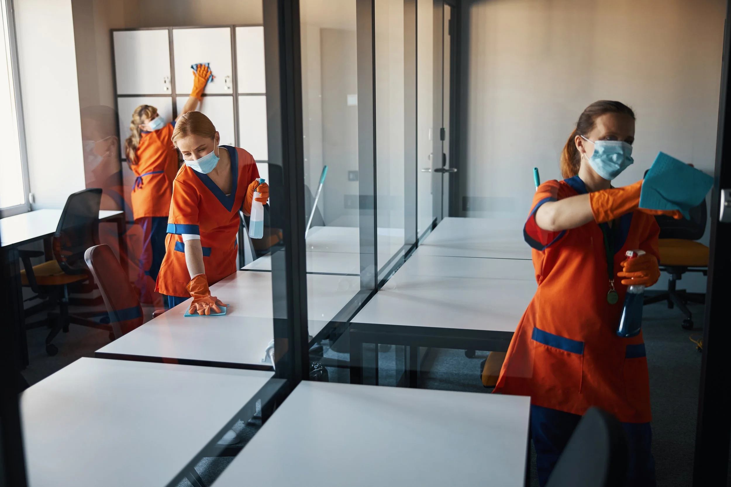 Cleaning staff wearing face masks, orange uniforms, and gloves, disinfecting office rooms with glass walls, using spray bottles and cleaning cloths.