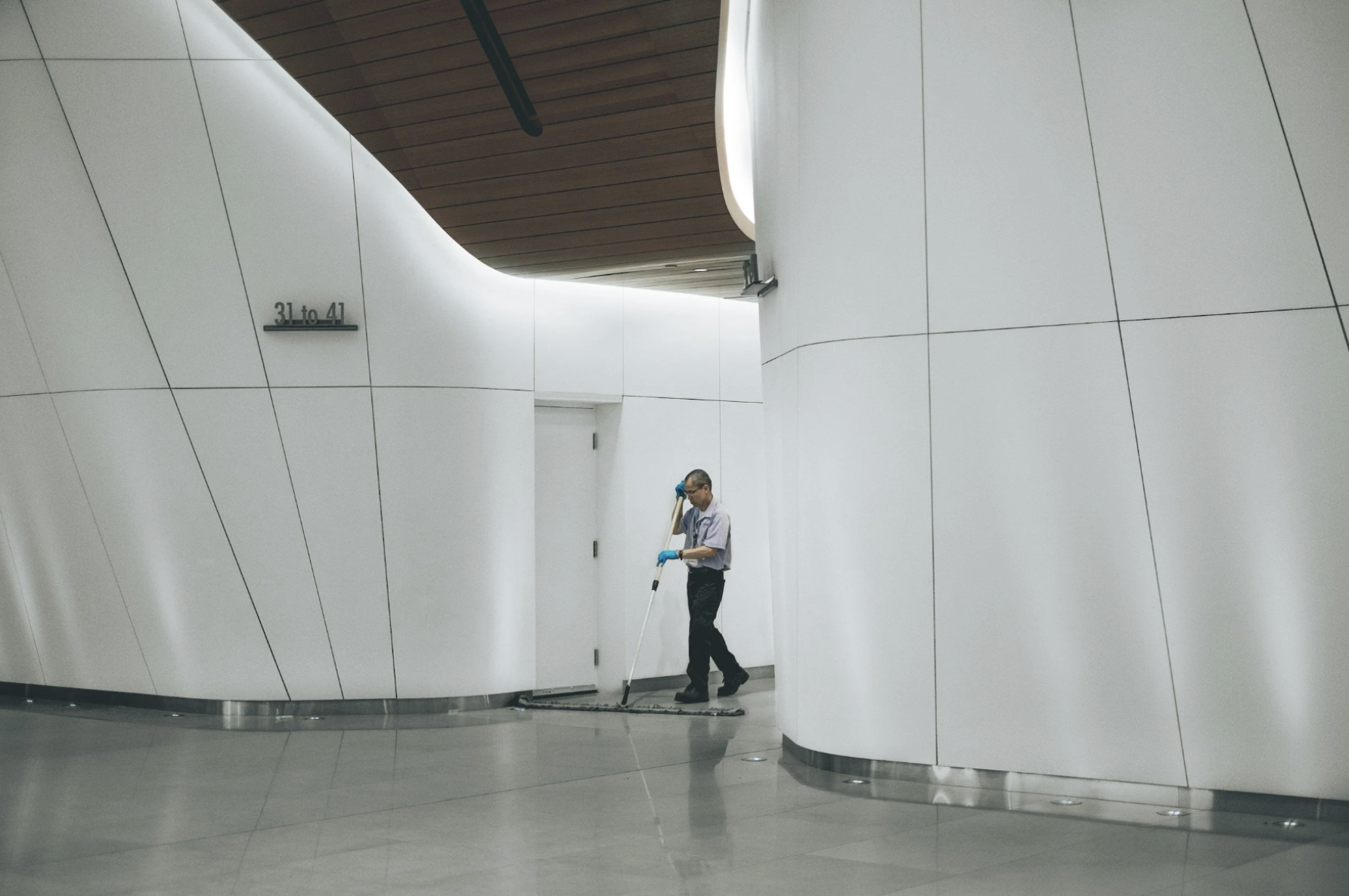 A man cleaning the floor with a mop in a modern, minimalistic interior space with white walls and wooden ceiling.