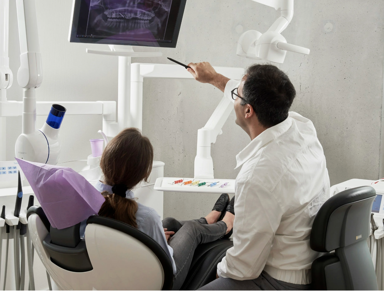 A young girl sitting in a dentist's chair with purple tissue paper on her head, talking to a dentist. The dentist is pointing at a monitor displaying a dental X-ray, while on a tray in front of the girl are dental models and colorful dental tools.