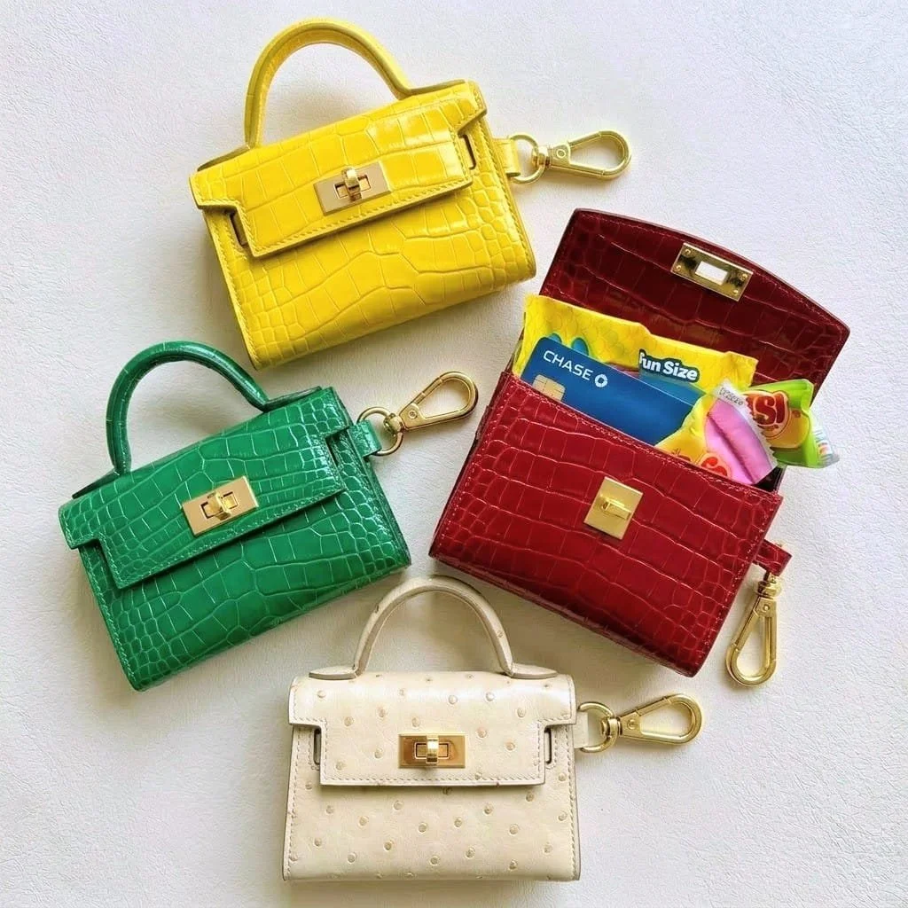 Luxurious colorful miniature keychain handbags in yellow, green, red, and white exotic-looking leather, with the red version open and emergency sugary snacks and a blue credit card, all with gold hardware and clips on a white surface.