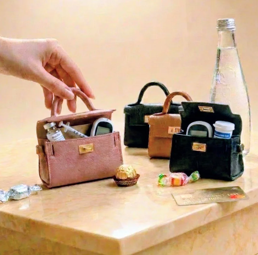 Leather miniature handbag keychains that can contain emergency insulin, glucose, and a credit card on a wooden table, shown next to a hand and a water bottle for size comparison.