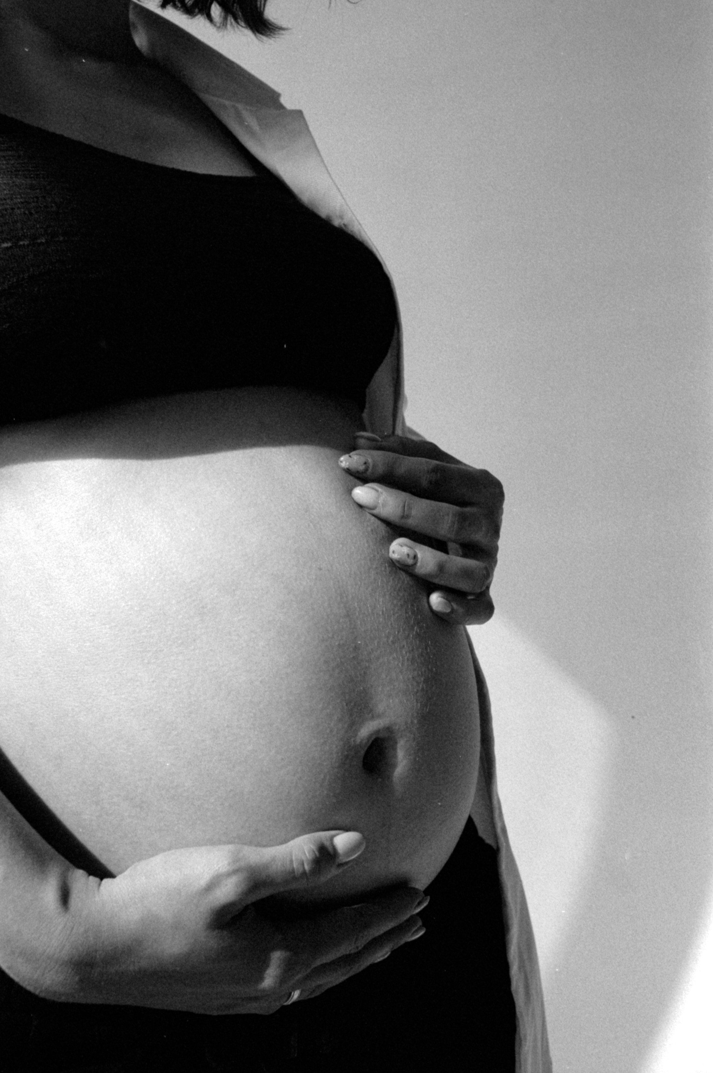 Black and white image of a person holding their pregnant belly with both hands. The person's face is not visible, and they are wearing a shirt with the sleeve rolled up.