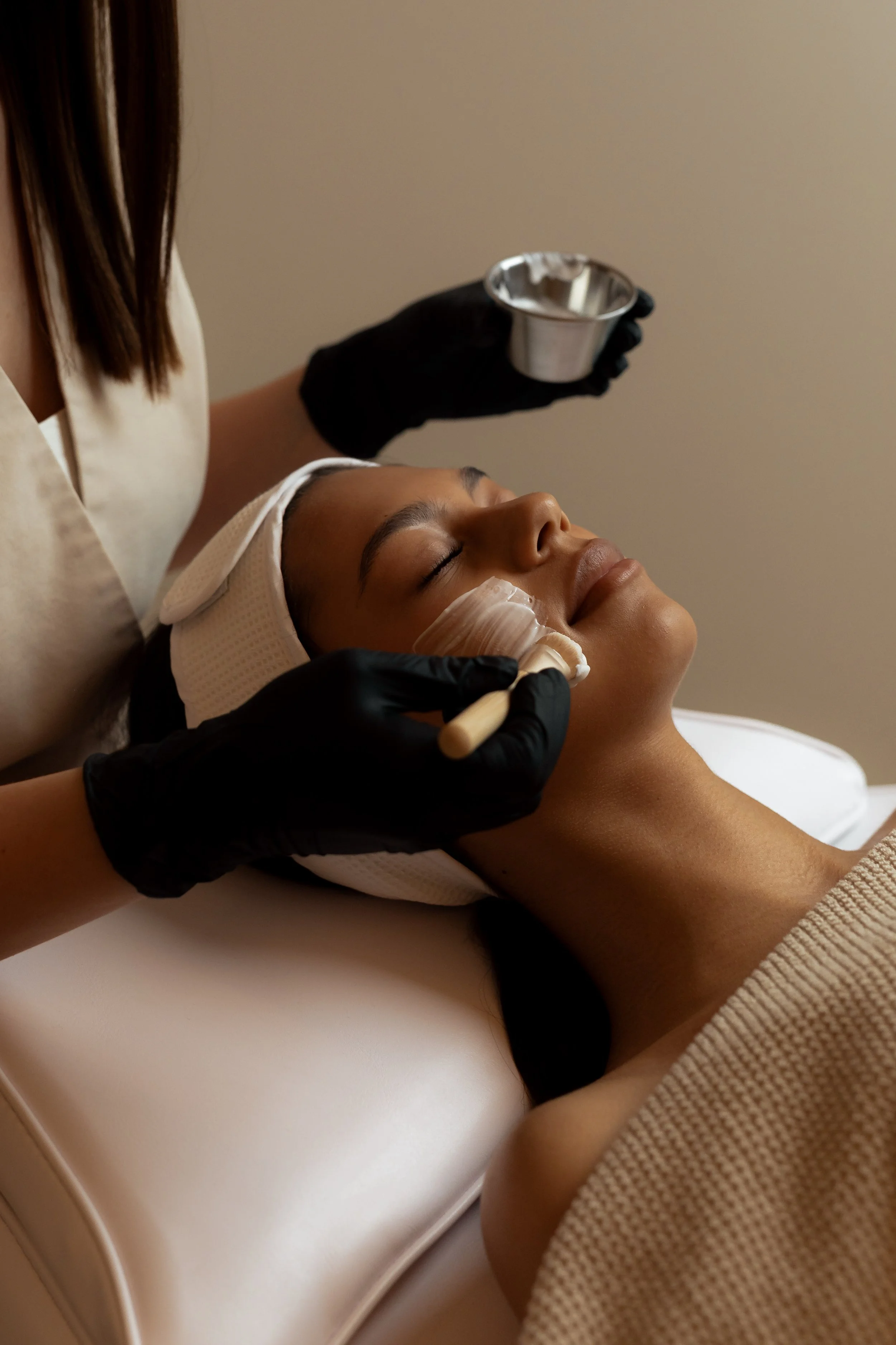 A woman receiving a facial treatment, lying down with a headband, while a professional wearing black gloves applies a product with a brush and holds a small metallic bowl.