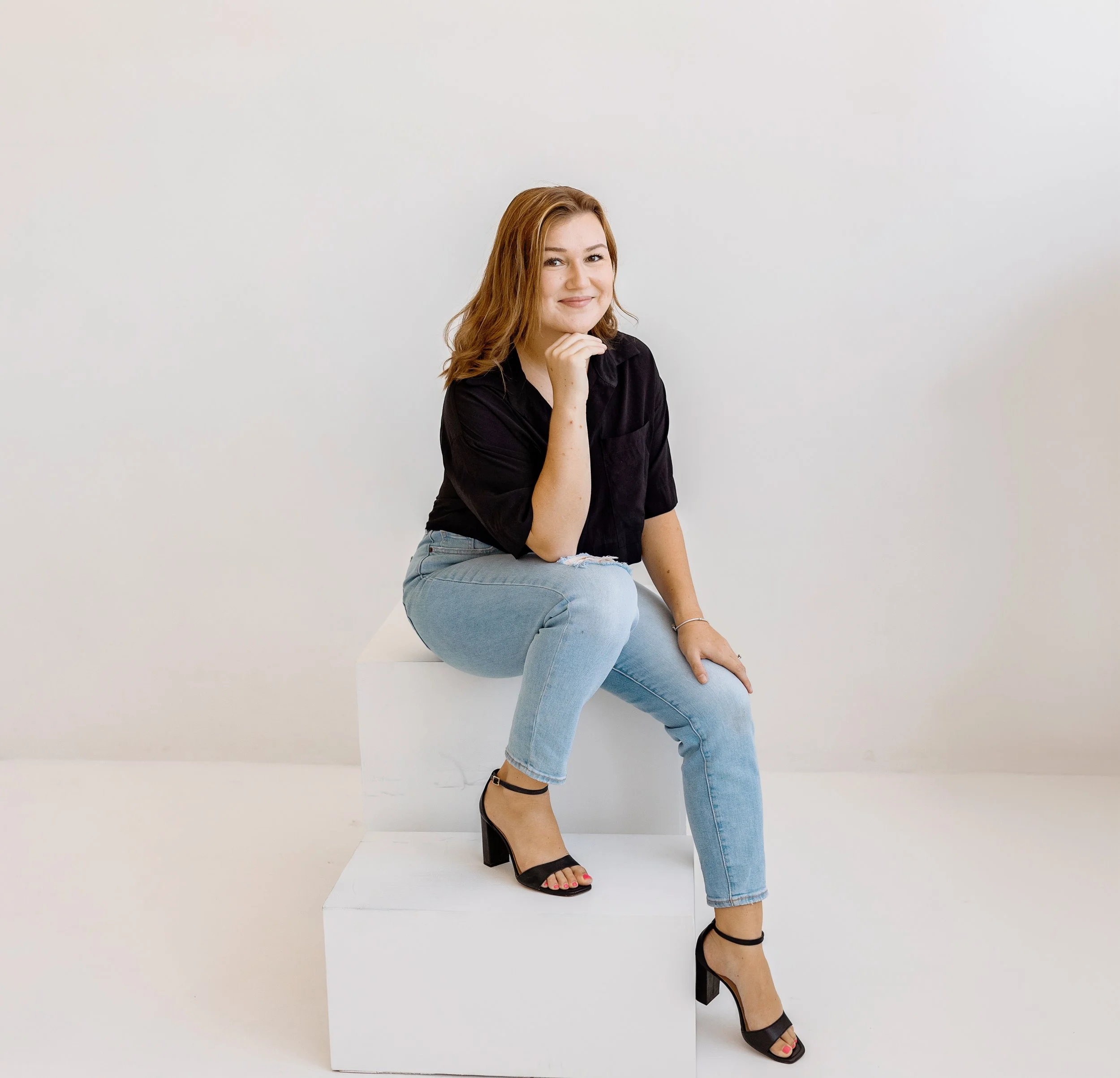 A woman with wavy auburn hair sitting on a white platform against a plain white background, wearing a black shirt, light blue jeans, and black high-heeled sandals.
