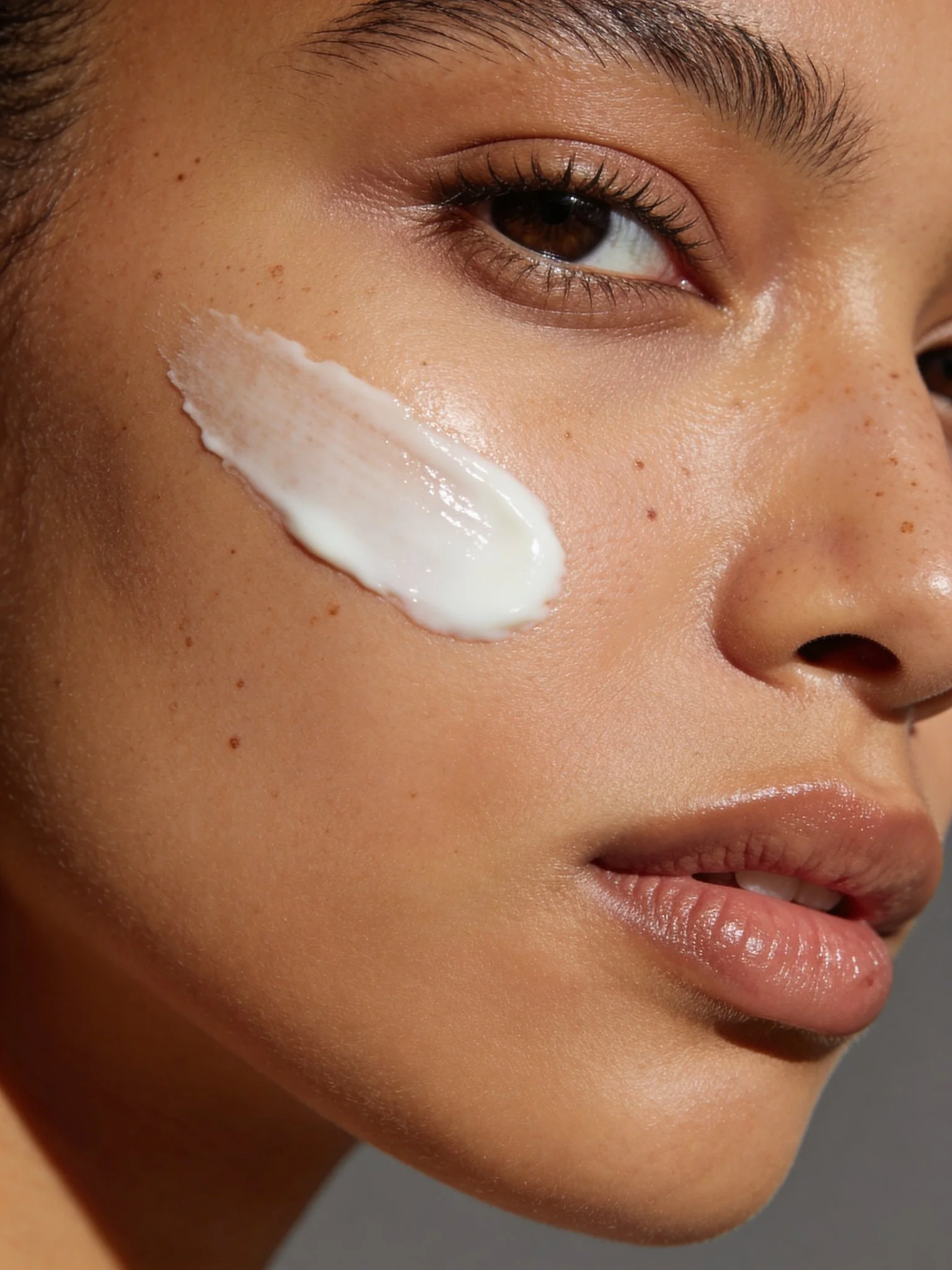 Close-up of a person's face with a dollop of white facial cream on their cheek, showing skin texture and facial features.