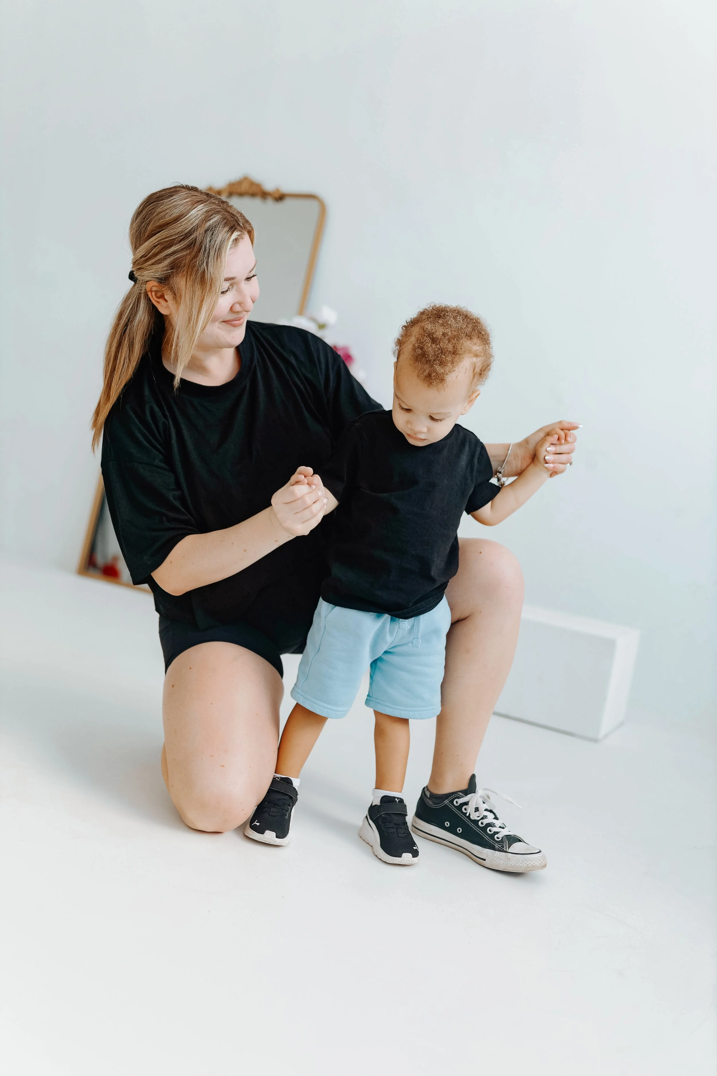 A woman with long blonde hair kneeling on one knee, holding the hands of a young boy with light curly hair. They are wearing black shirts, and the boy is in light blue shorts and black sneakers. The woman is smiling softly, and the boy looks downward. The background is plain white with a mirror and some decor blurred behind them.