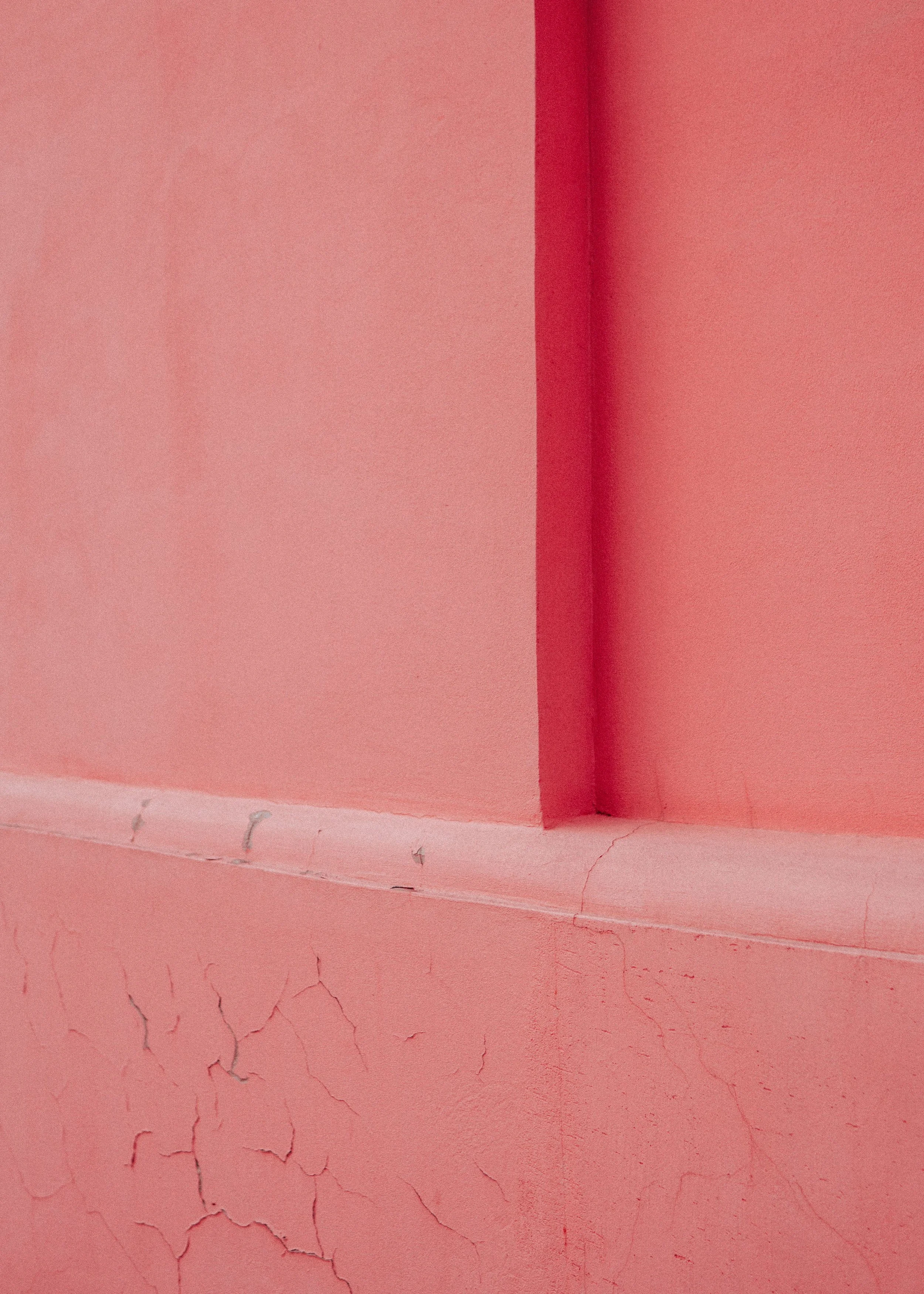 Close-up of a pink wall with visible cracks and uneven paint, showing a corner with a ledge and some damage.