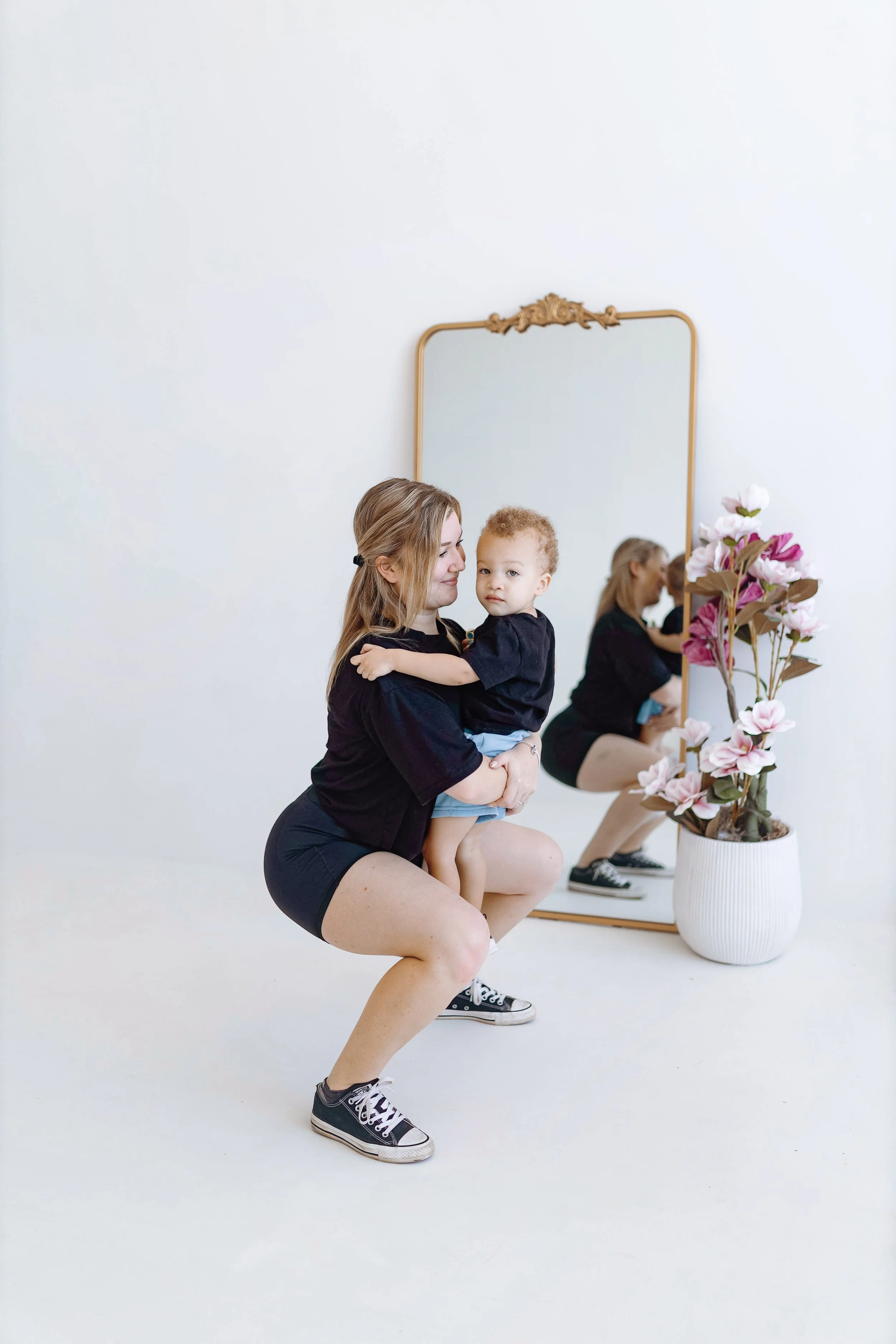 A woman in black athletic apparel and sneakers squatting, holding a young child in black shirt and shorts, in front of a large mirror with a decorative gold frame. The mirror reflects a woman sitting on a bench, with pink and white flowers in a large white vase nearby.