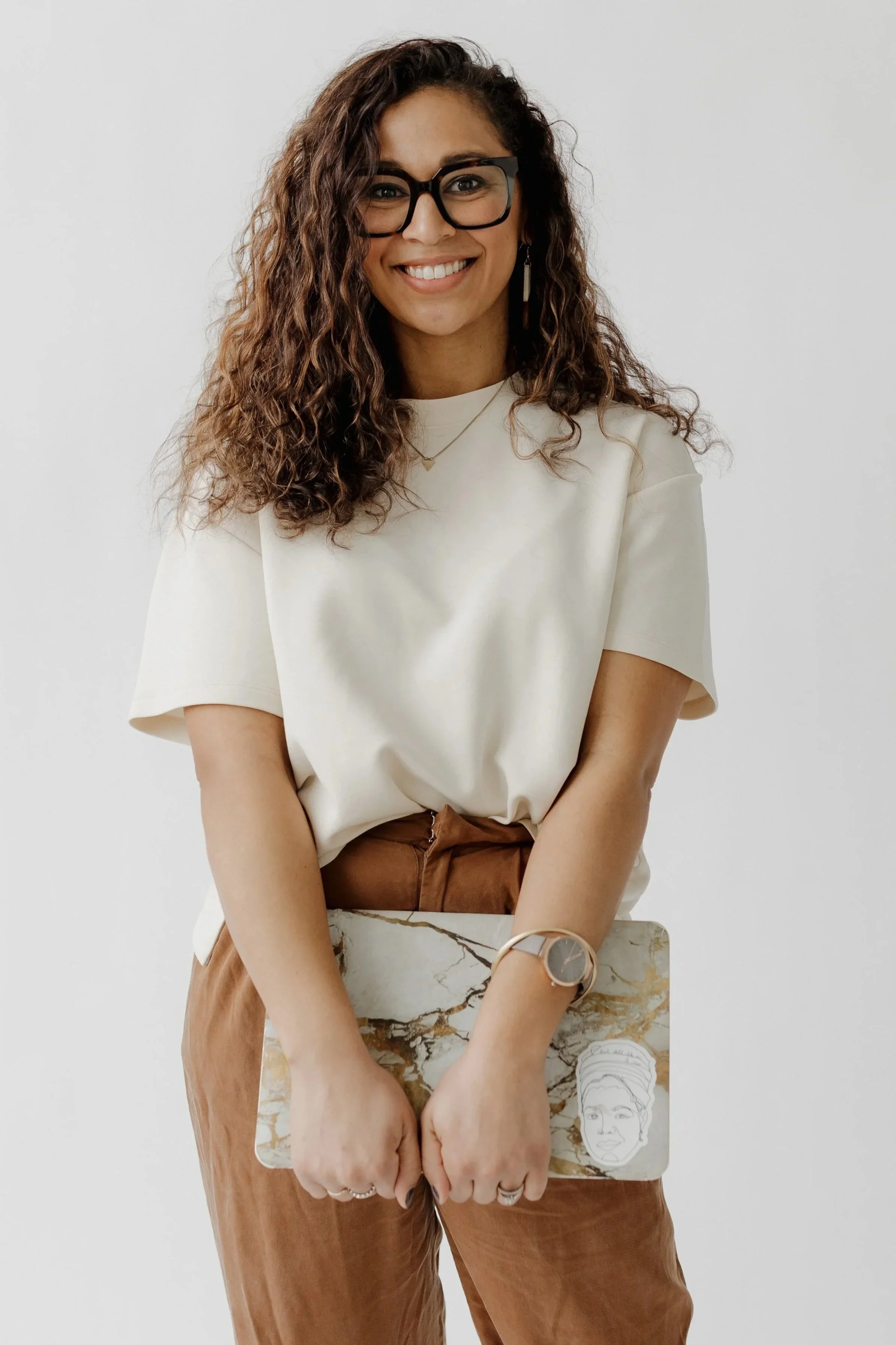 A woman with curly brown hair wearing glasses, a white t-shirt, and brown pants, smiling and holding a marble-patterned laptop with a face sketch on it, standing against a plain white wall.