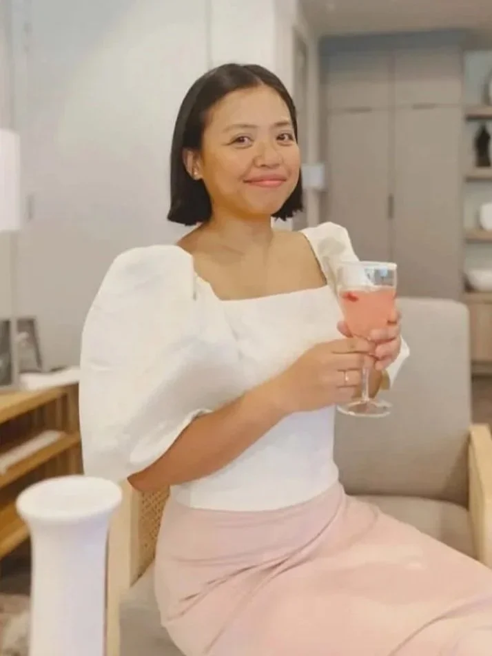 A woman with short black hair and light skin, wearing a white puff-sleeve top and beige skirt, sitting and holding a glass of pink drink, smiling in a cozy indoor setting.