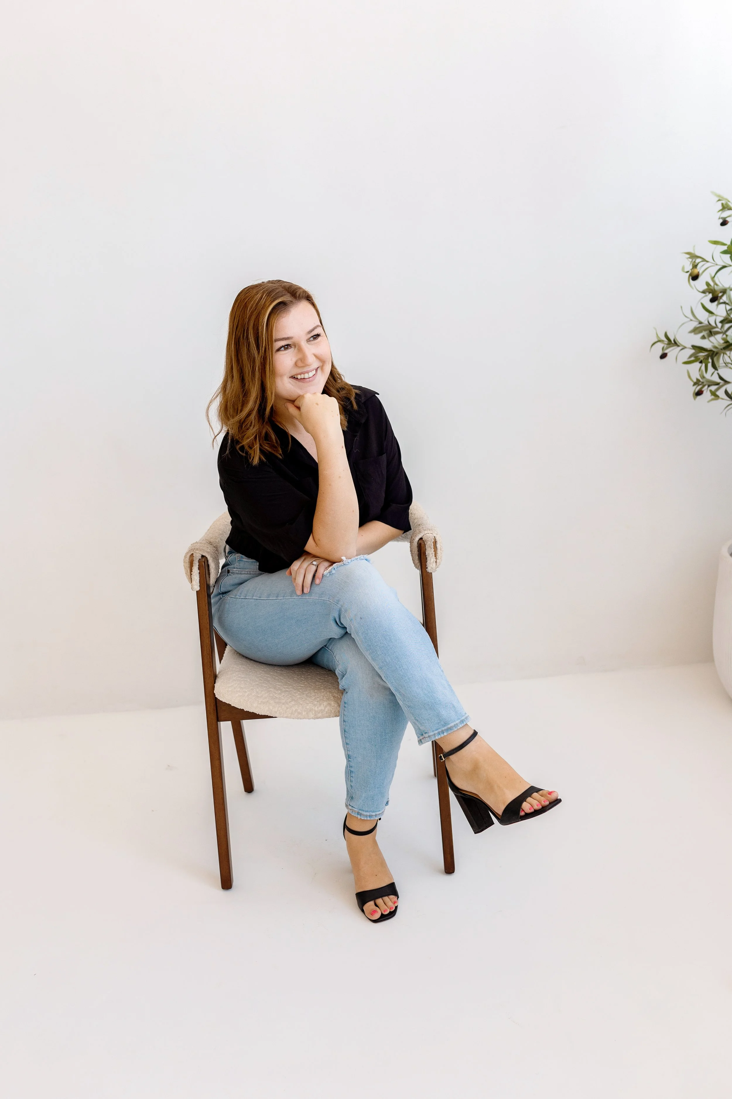 A young woman with reddish-brown hair, smiling and sitting on a wooden chair with a cushioned seat, in front of a plain white wall. She is wearing a black top, light blue jeans, and black high-heeled sandals, with one leg crossed over the other.