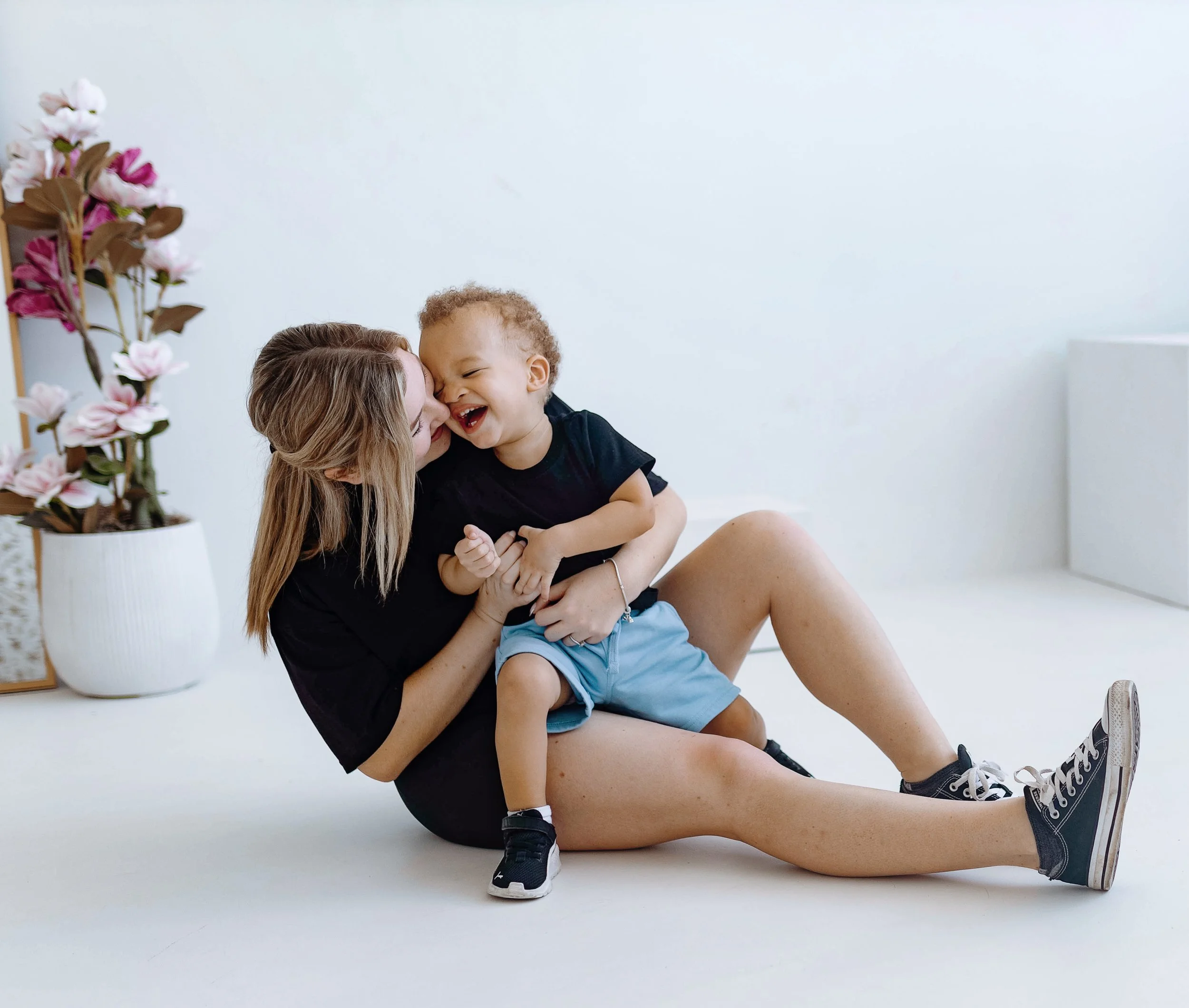 A woman with long blonde hair holding a young boy with curly hair in her arms, both smiling and laughing. They are positioned against a plain white background with a potted plant with pink and white flowers on the left side of the image.