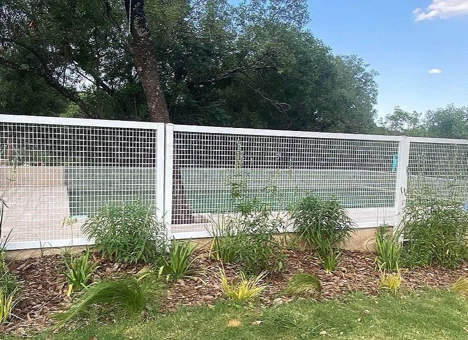 Cerca blanca de metal con plantas y arbustos en primer plano, árboles y cielo azul con nubes al fondo.